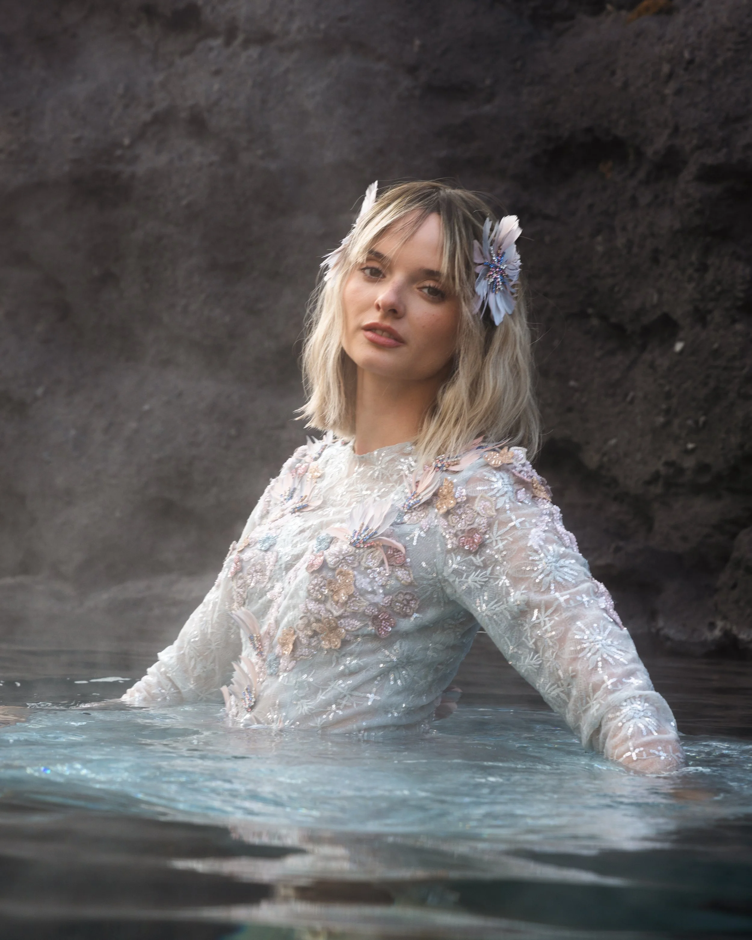 A woman with blonde hair and flowers in her hair, wearing a white embroidered dress, standing in water near a rocky shoreline.