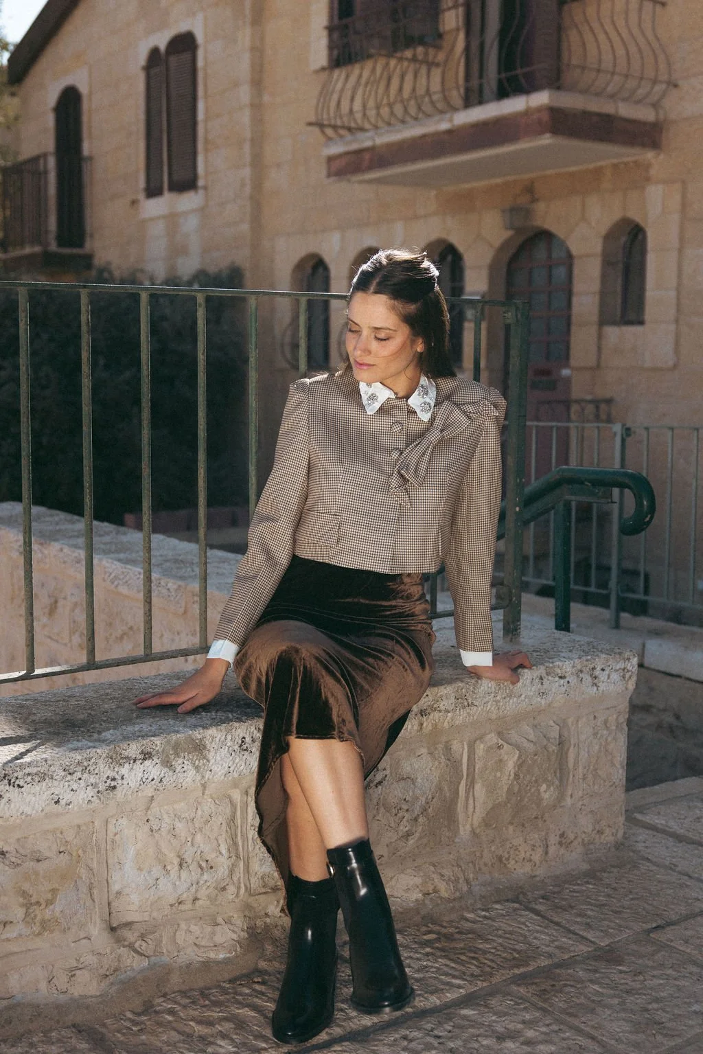 A woman sitting on a stone ledge outside, wearing a checkered blazer with ruffled collar, velvet skirt, and black ankle boots.