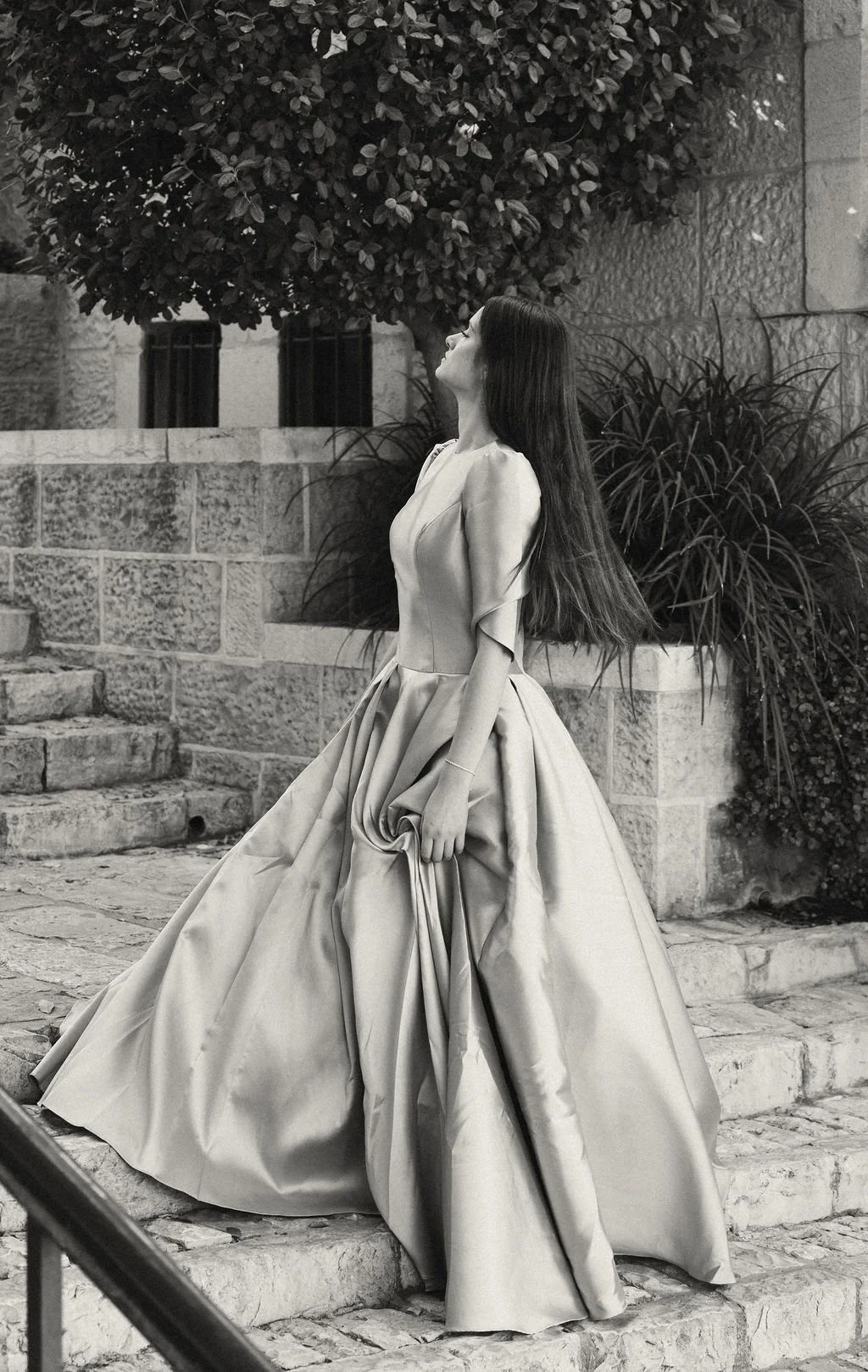 A woman in an elegant, flowing gown walking down stone steps outdoors, with bushes and a tree in the background, captured in black and white.