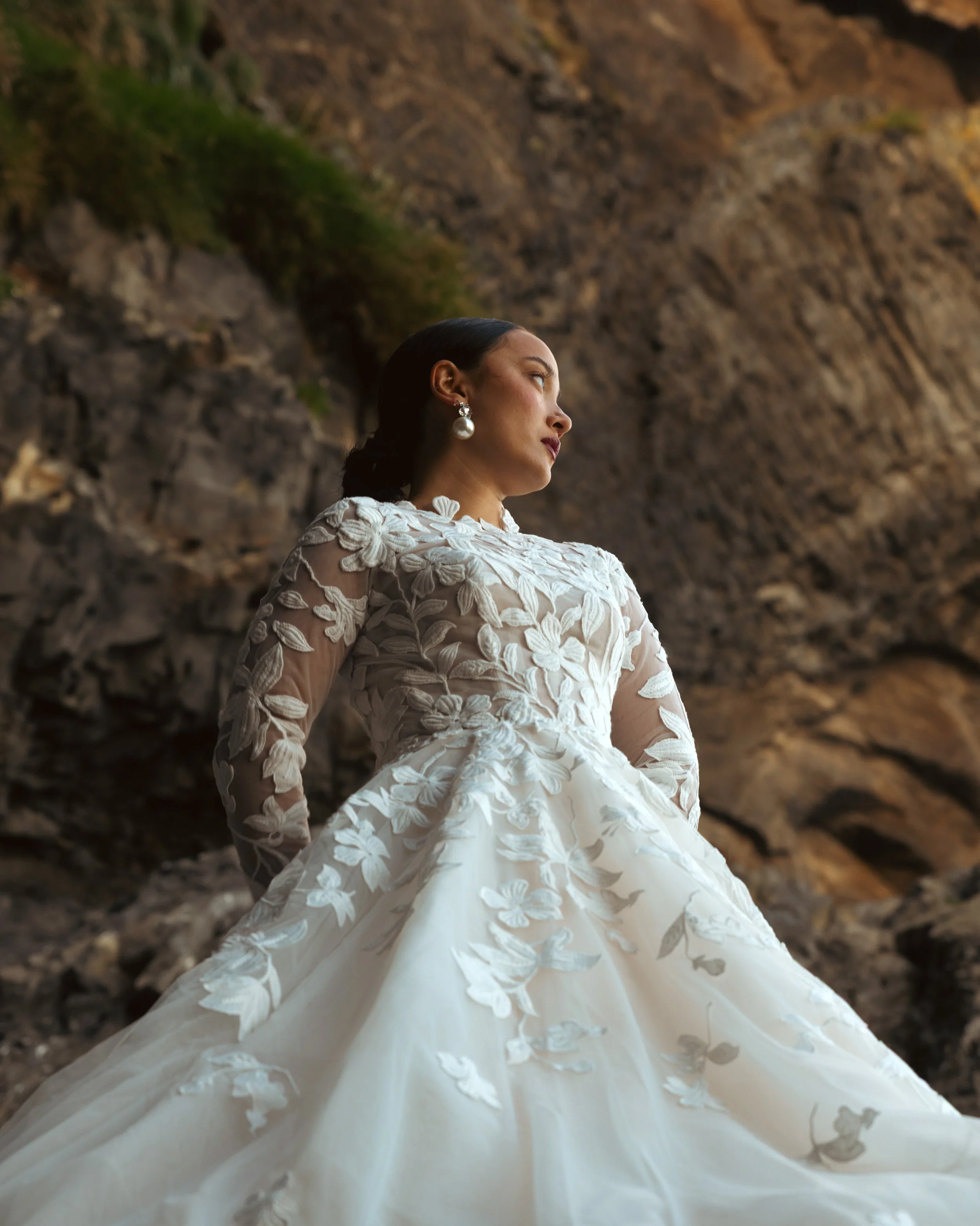 A woman in a white wedding dress with floral embroidery stands outdoors against a rocky background, looking to the side with a serious expression.