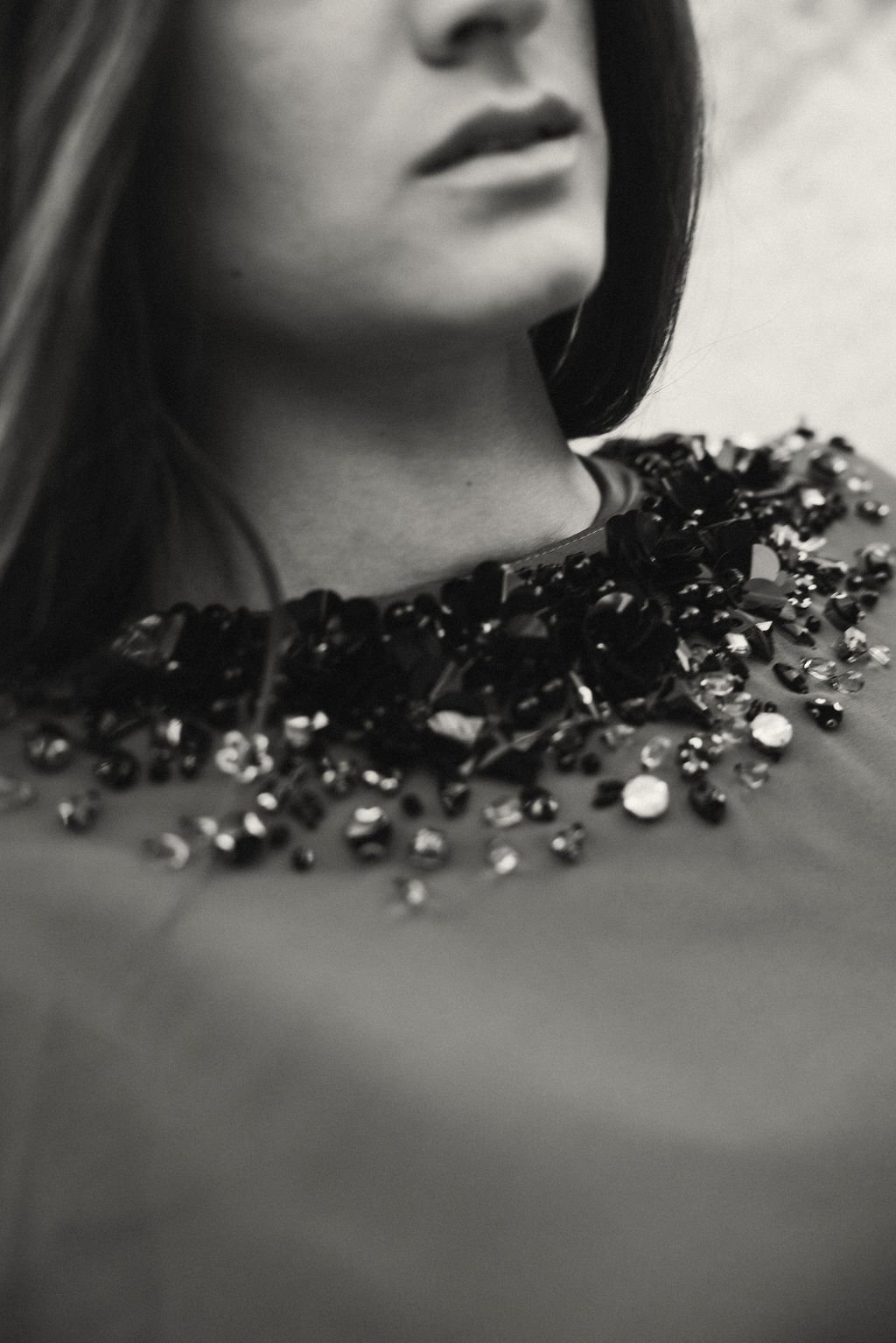 Close-up black and white photo of a woman's face and neck, adorned with a bejeweled neckline, with her lips slightly parted and dark hair partially covering her face.