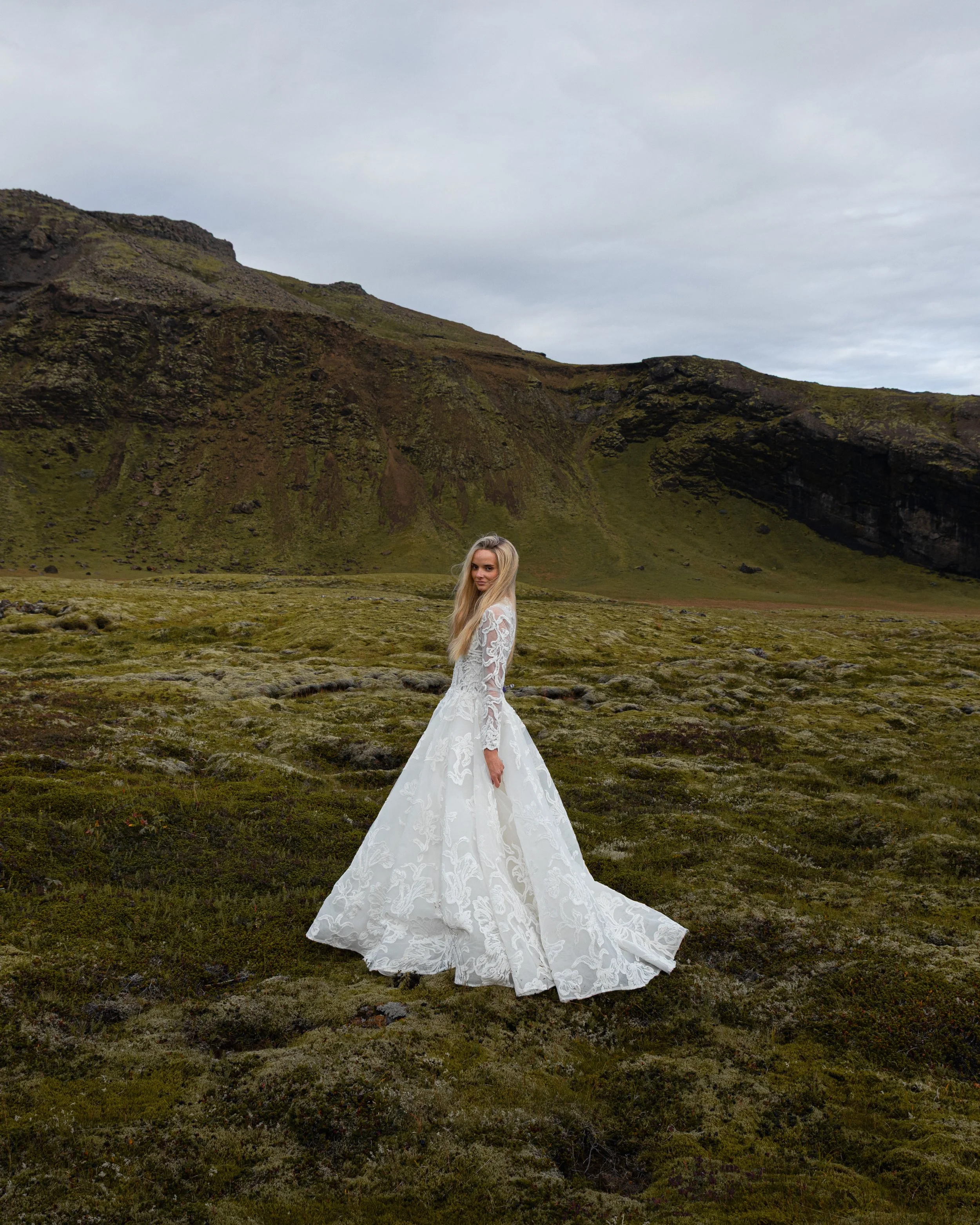 A woman in a long, white lace wedding dress stands on a grassy plain with moss and rocks, with a mountain landscape and cloudy sky in the background.