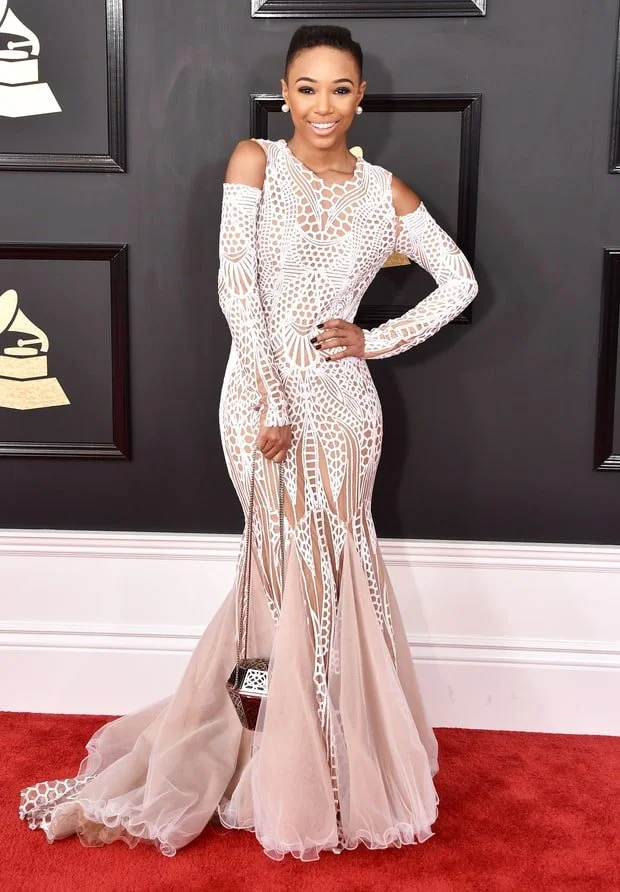 Woman in a white, intricately patterned sheer gown with cut-out details, standing on a red carpet at an awards event.