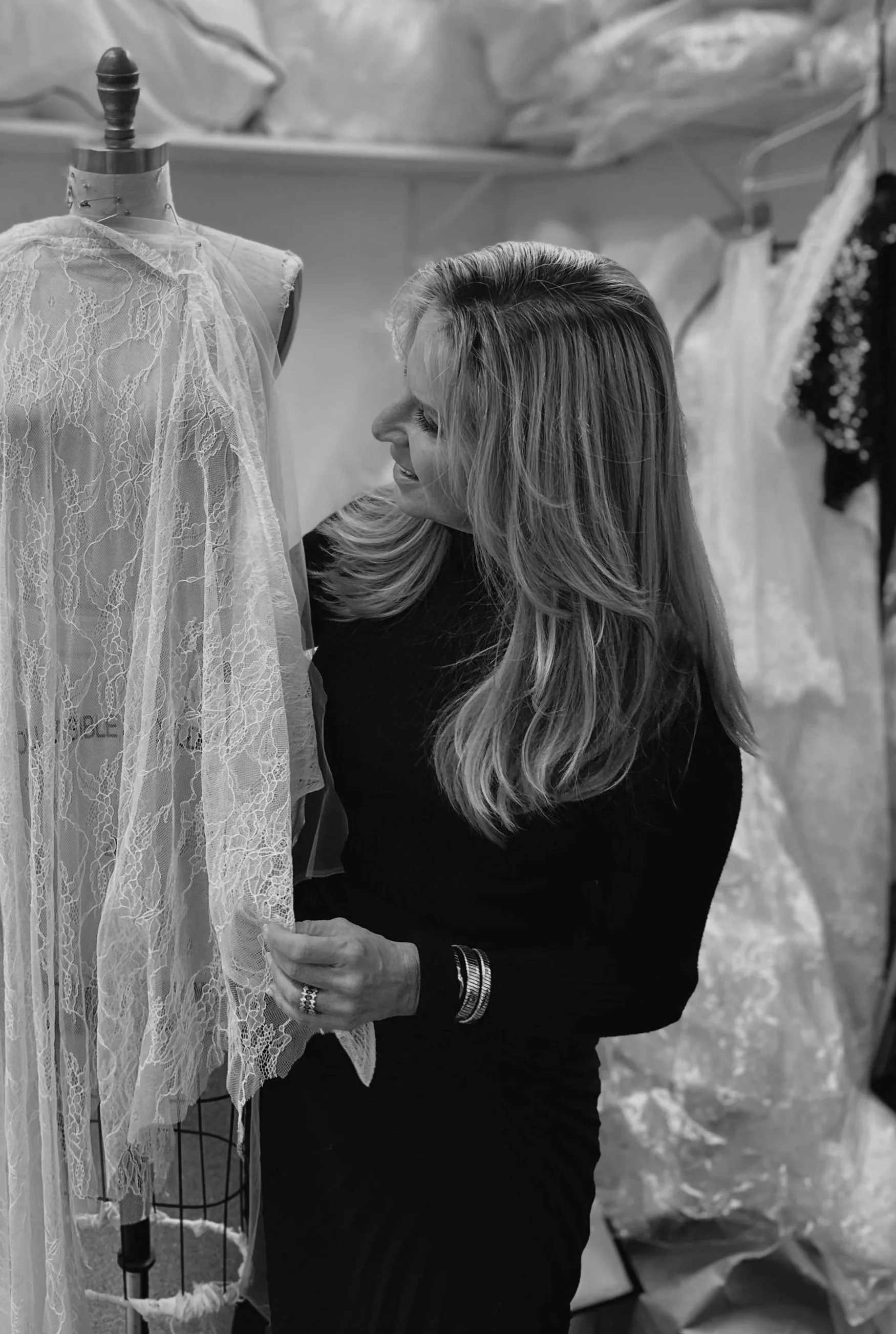 A woman with long blonde hair smiling and looking at a lace wedding dress on a mannequin in a bridal shop.