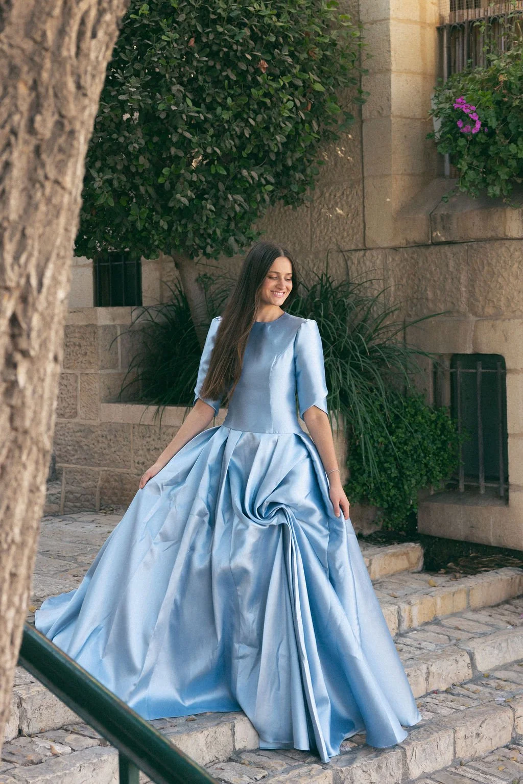 A woman wearing a long, elegant blue satin gown with puffed sleeves and a full skirt, standing outdoors near stone steps and greenery, smiling happily.