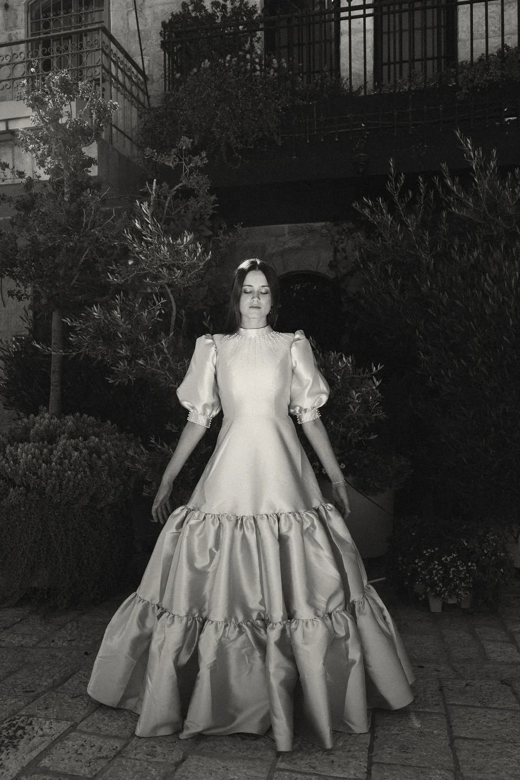 A woman in a vintage long satin dress with puffed sleeves is standing outdoors at night with her eyes closed. There are plants and a building with a balcony behind her in black and white.