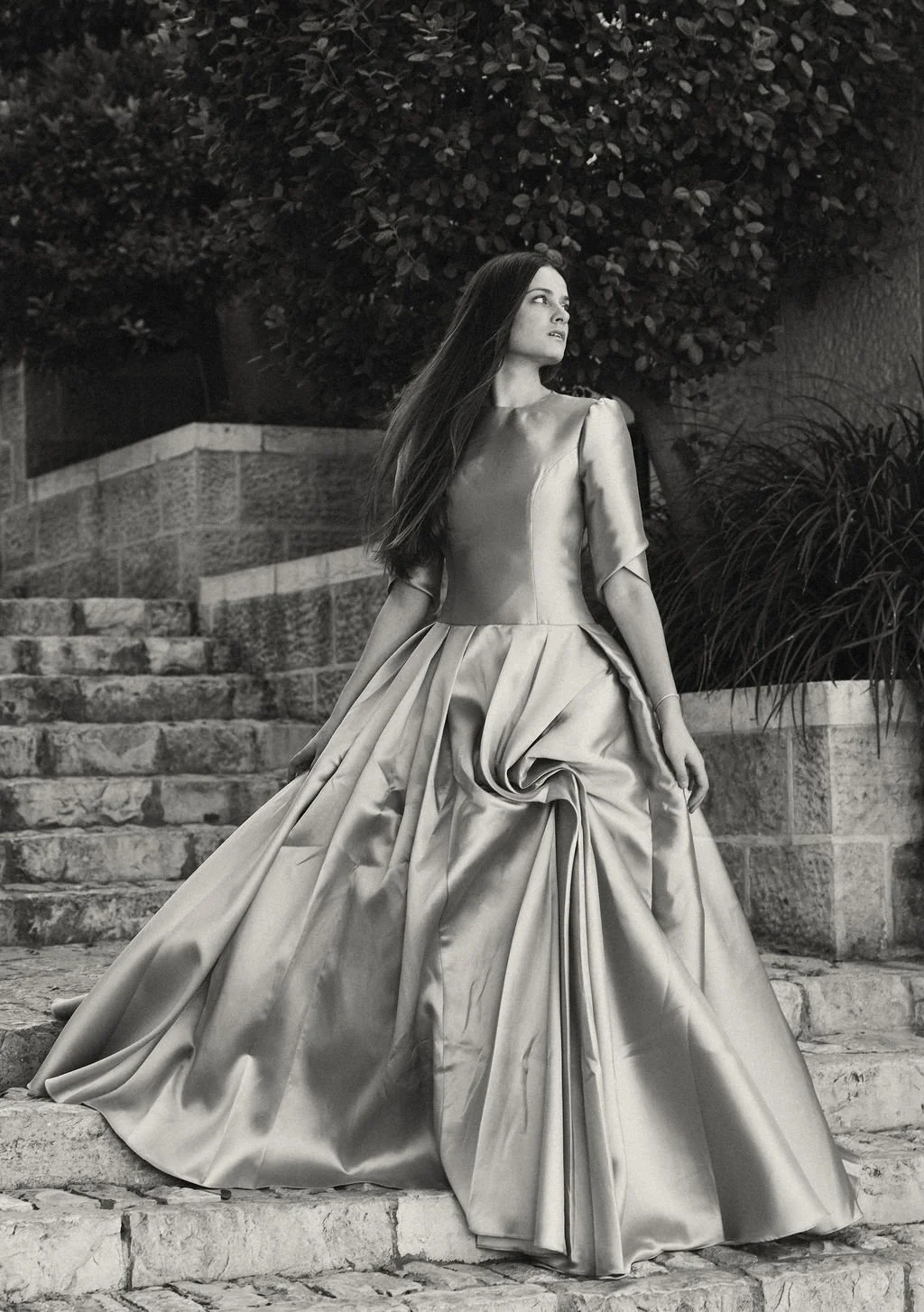 A woman in an elegant, flowing gown standing on stone stairs outdoors at night, with trees and shrubbery in the background.