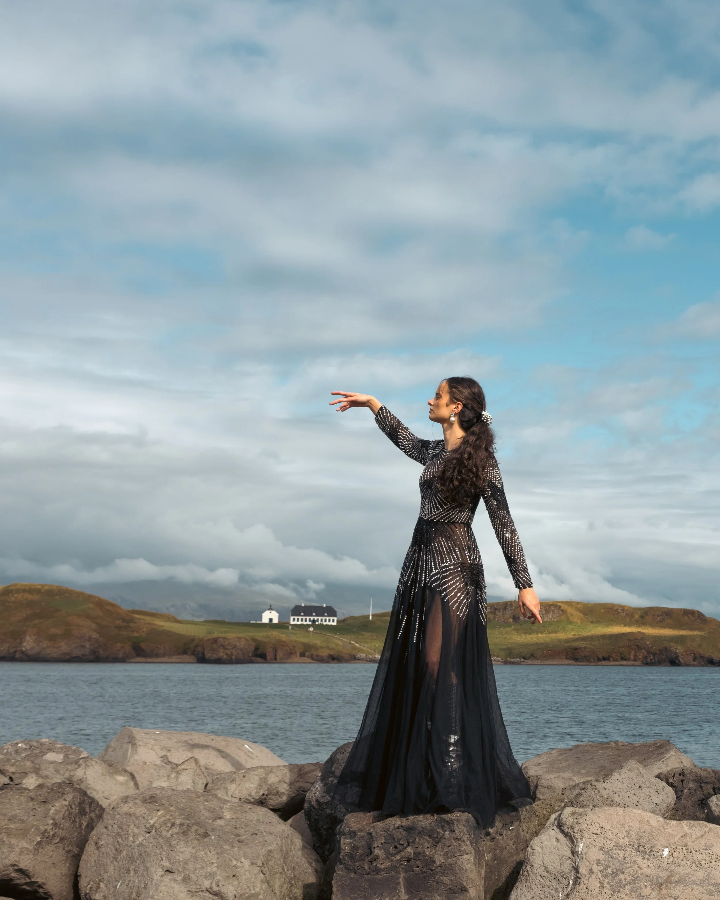 A woman in a black, sequined, floor-length dress standing on rocks by the water, with her right arm extended outward, against a backdrop of a cloudy sky and distant houses.