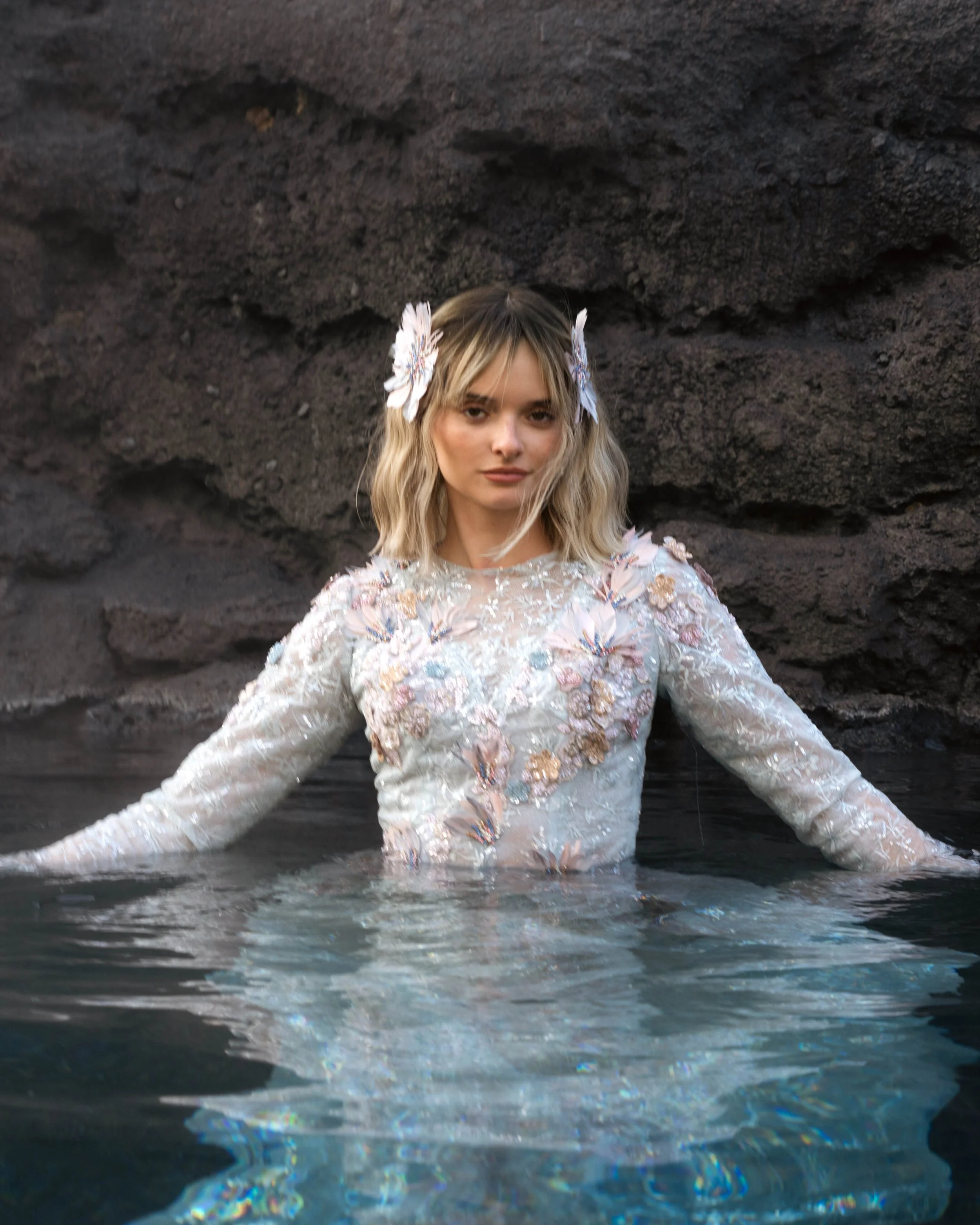 A woman with blonde hair and butterfly hair clips wears a floral embroidered long sleeve dress and stands in water against a rocky backdrop.