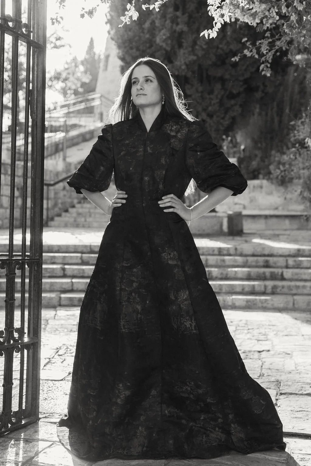 A woman in a dark, full-length dress standing outdoors with hands on her hips near a gate and stone steps, with trees and sunlight in the background.
