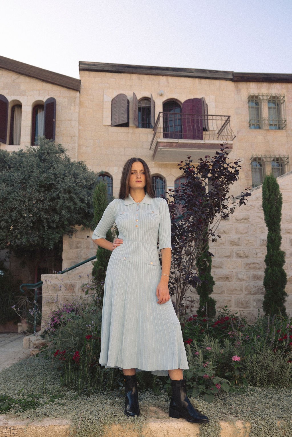 A woman in a light-colored, long pleated dress and black boots standing outdoors in front of a stone building with plants and shrubs.