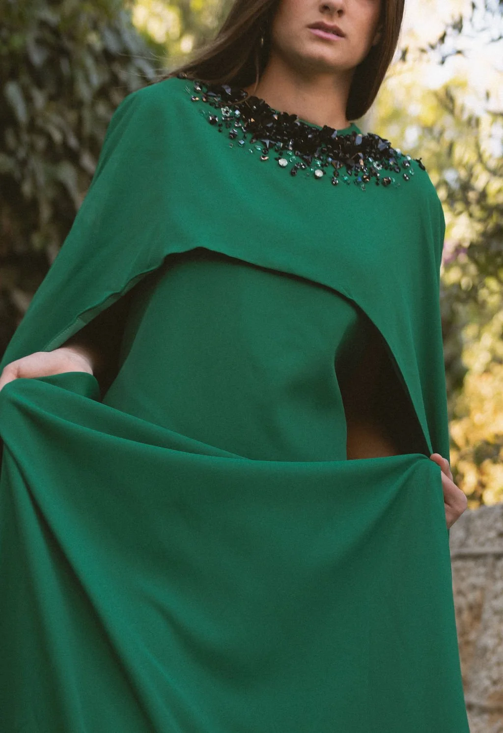 A woman outdoors wearing a green dress with black embellishments around the neckline, holding the hem of her dress. Background features trees and foliage.