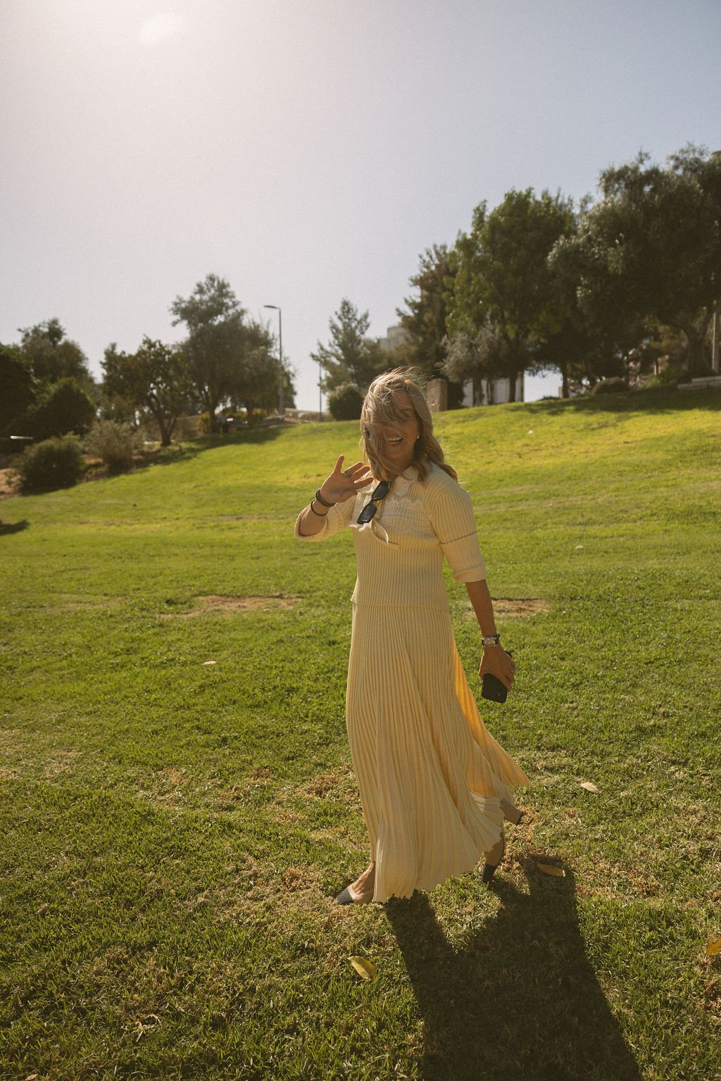A woman in a yellow pleated dress is standing on a grassy park during daytime, smiling and waving with one hand while holding a phone in the other. There are trees and a slight hill in the background.
