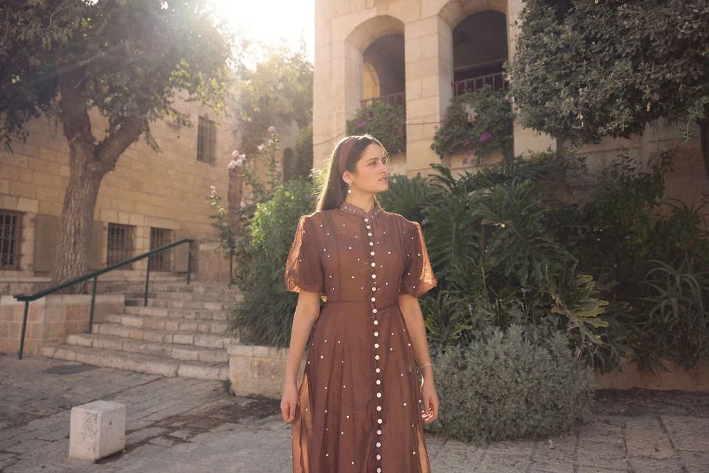 A woman in a brown dress with pearl accents standing outdoors near stone buildings and greenery, with sunlight filtering through trees.