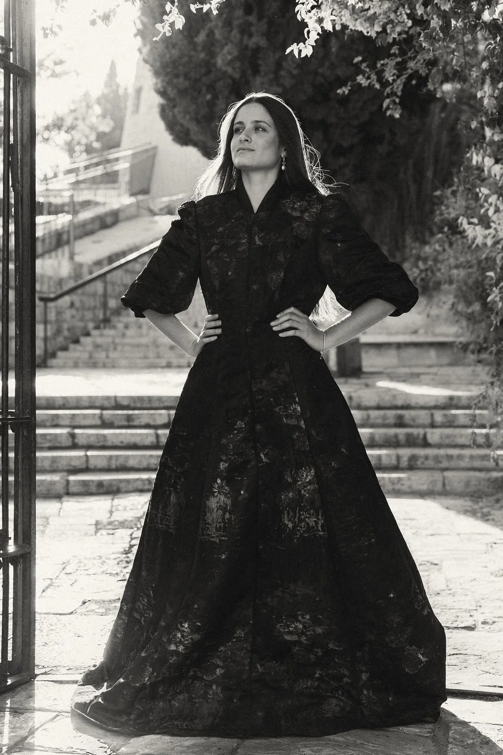A woman in a long black dress stands outdoors, hands on her hips, with trees and stairs in the background, captured in black and white.