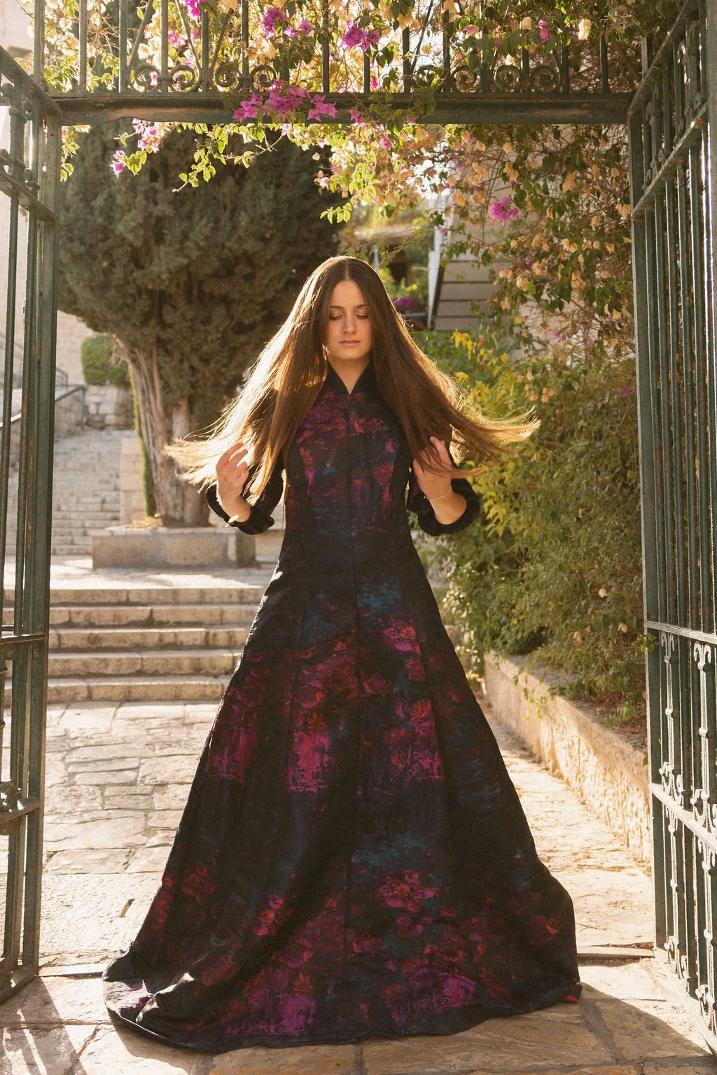 A woman stands in an open gate, with her eyes closed and hair flowing, wearing a long, dark, floral dress. The background includes stone stairs, trees, and flowering bushes, illuminated by warm sunlight.