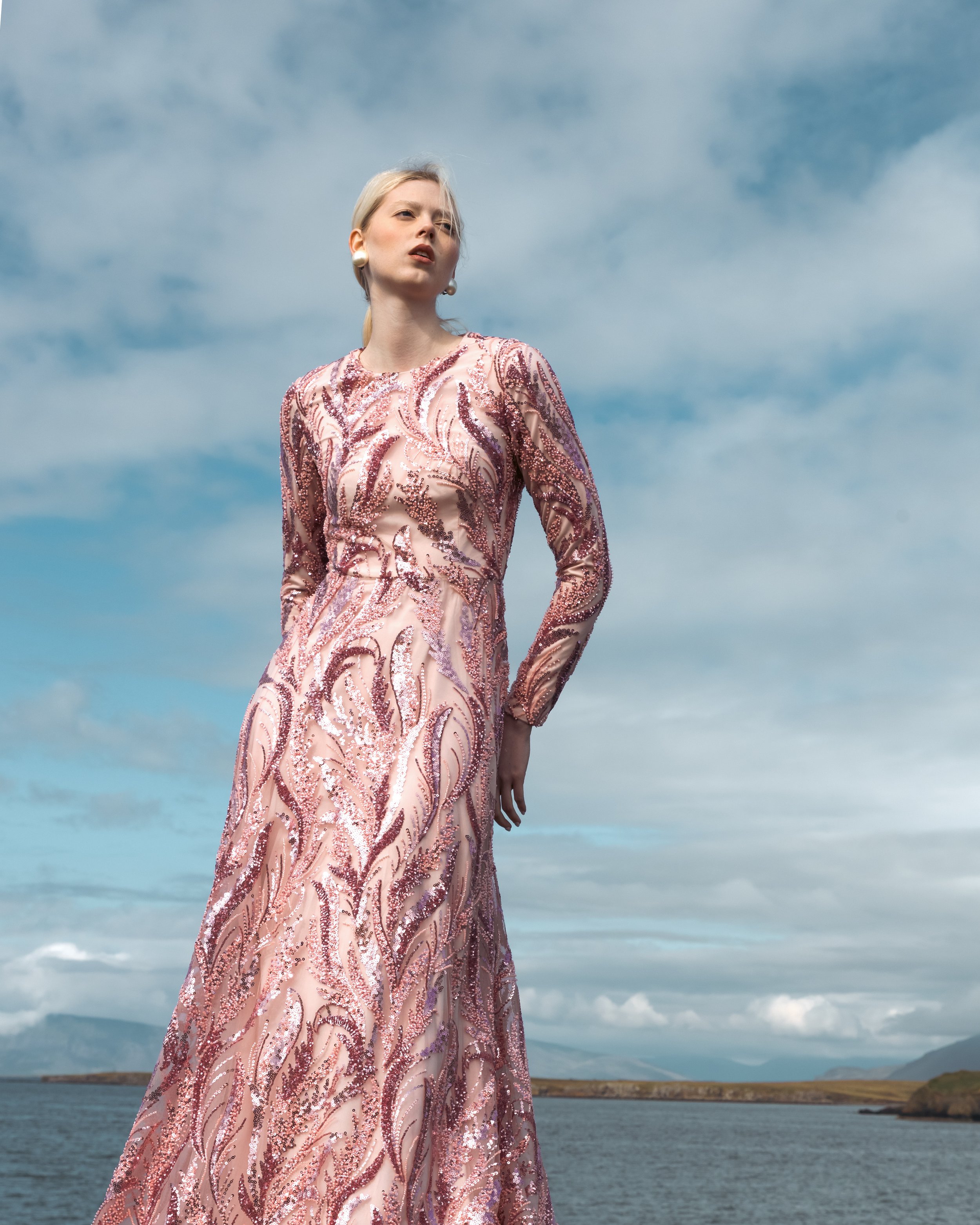 A woman with blonde hair in a pink, sequined, embroidered gown standing outdoors near a body of water under a blue sky with clouds.