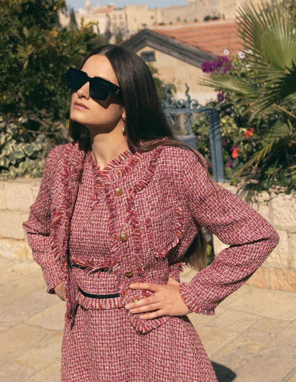 A woman with long dark hair wearing large black sunglasses and a pink tweed jacket with gold buttons, posing outdoors with her hand on her hip, in front of a garden background with plants and buildings.