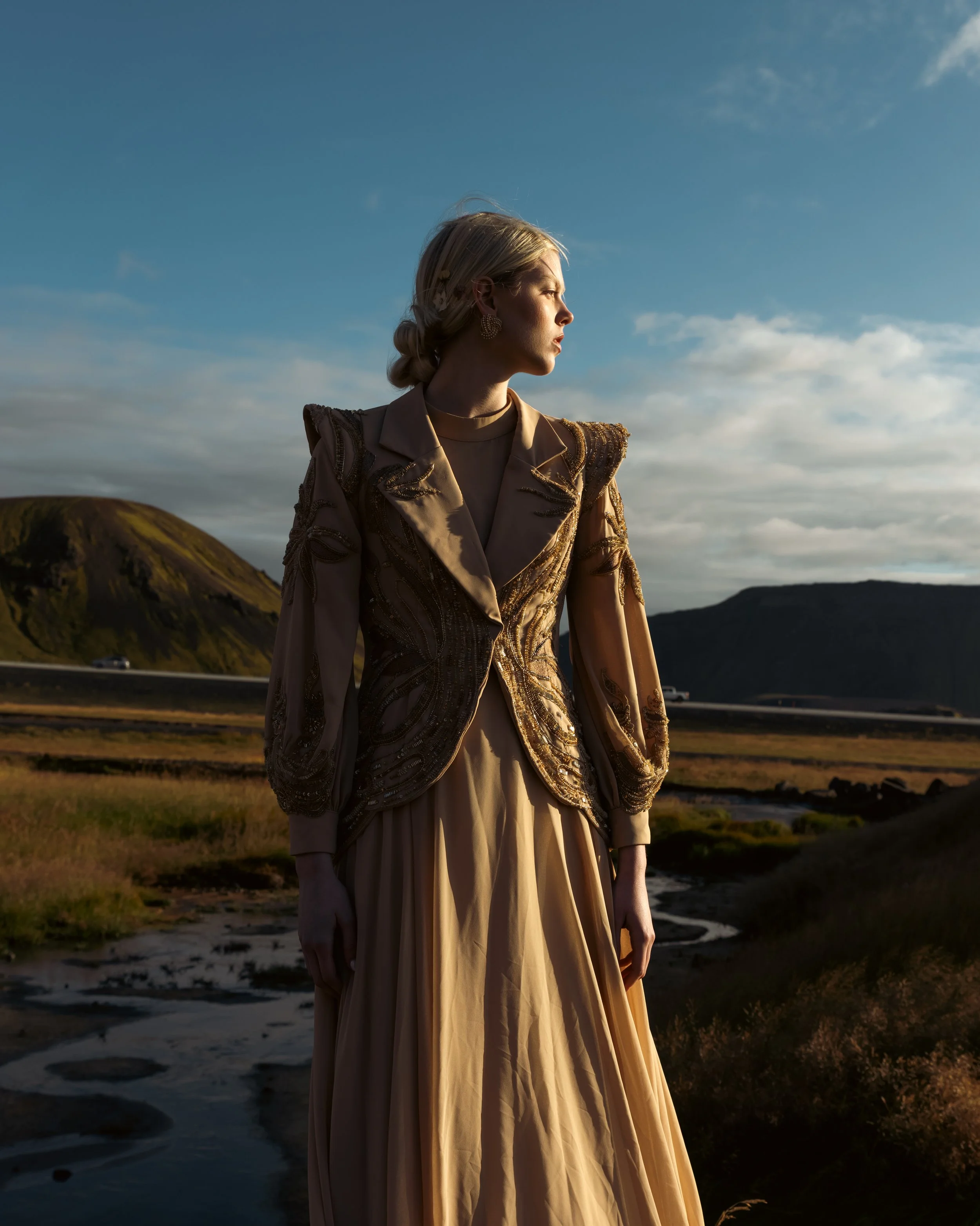 A woman standing outdoors in a beige and gold embroidered jacket and long beige skirt, with mountains and a stream in the background during sunset.