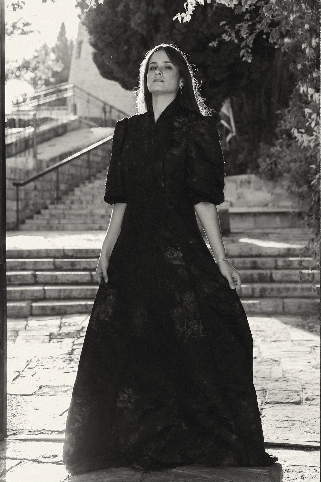 A woman in a long, black lace dress standing outdoors on stone steps, with trees and sunlight in the background.