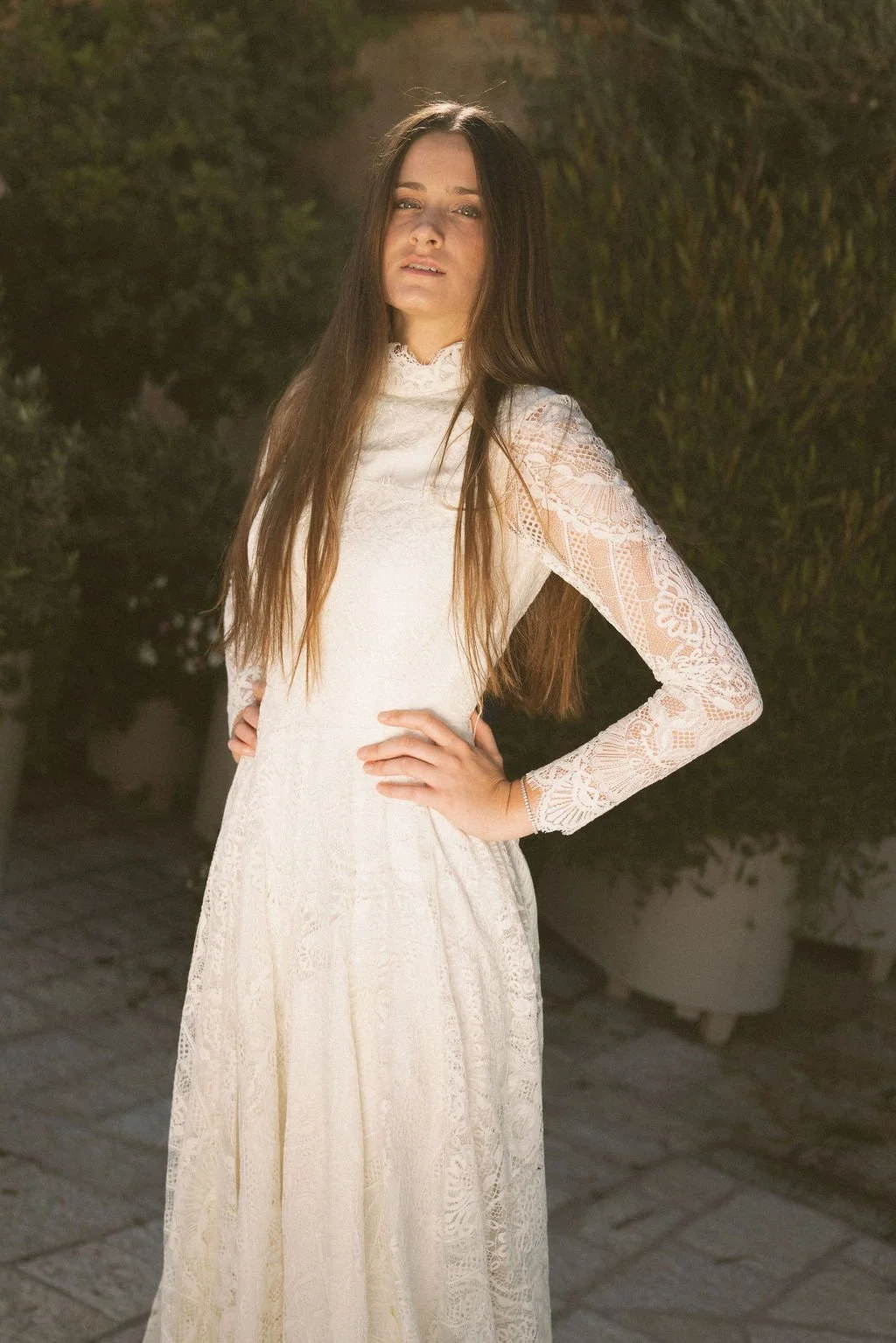 Young woman with long brown hair wearing a white lace dress, posing outdoors with hands on hips and a serious expression.