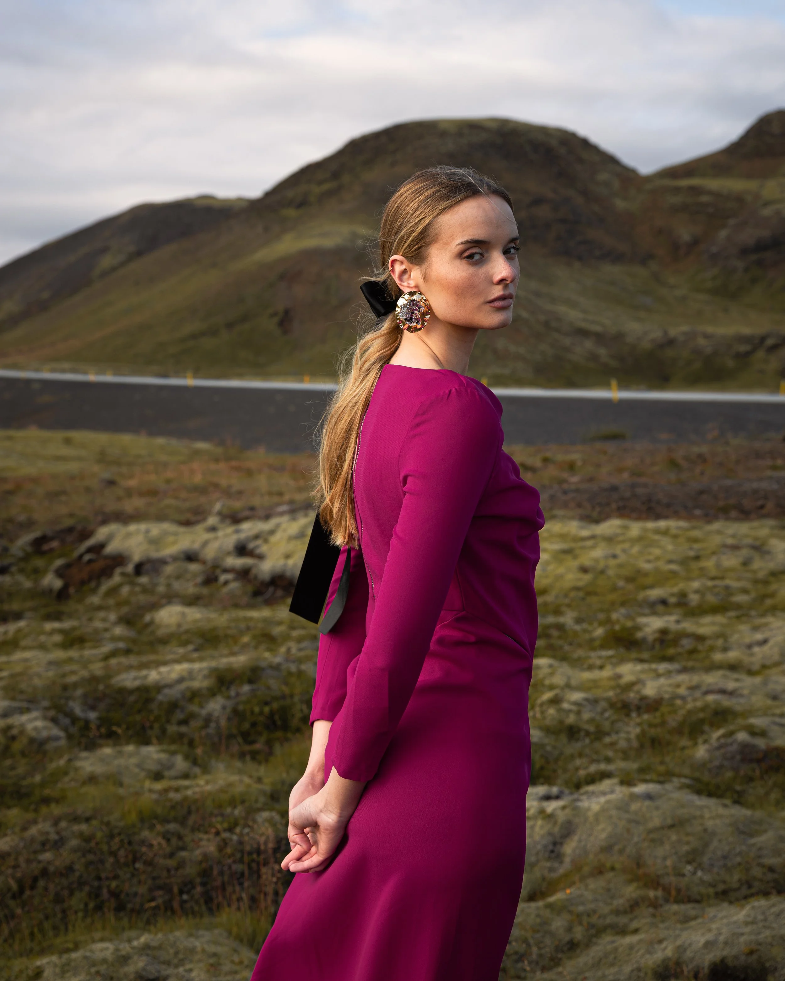 A woman wearing a magenta dress and colorful earrings stands outdoors in a mountainous landscape.