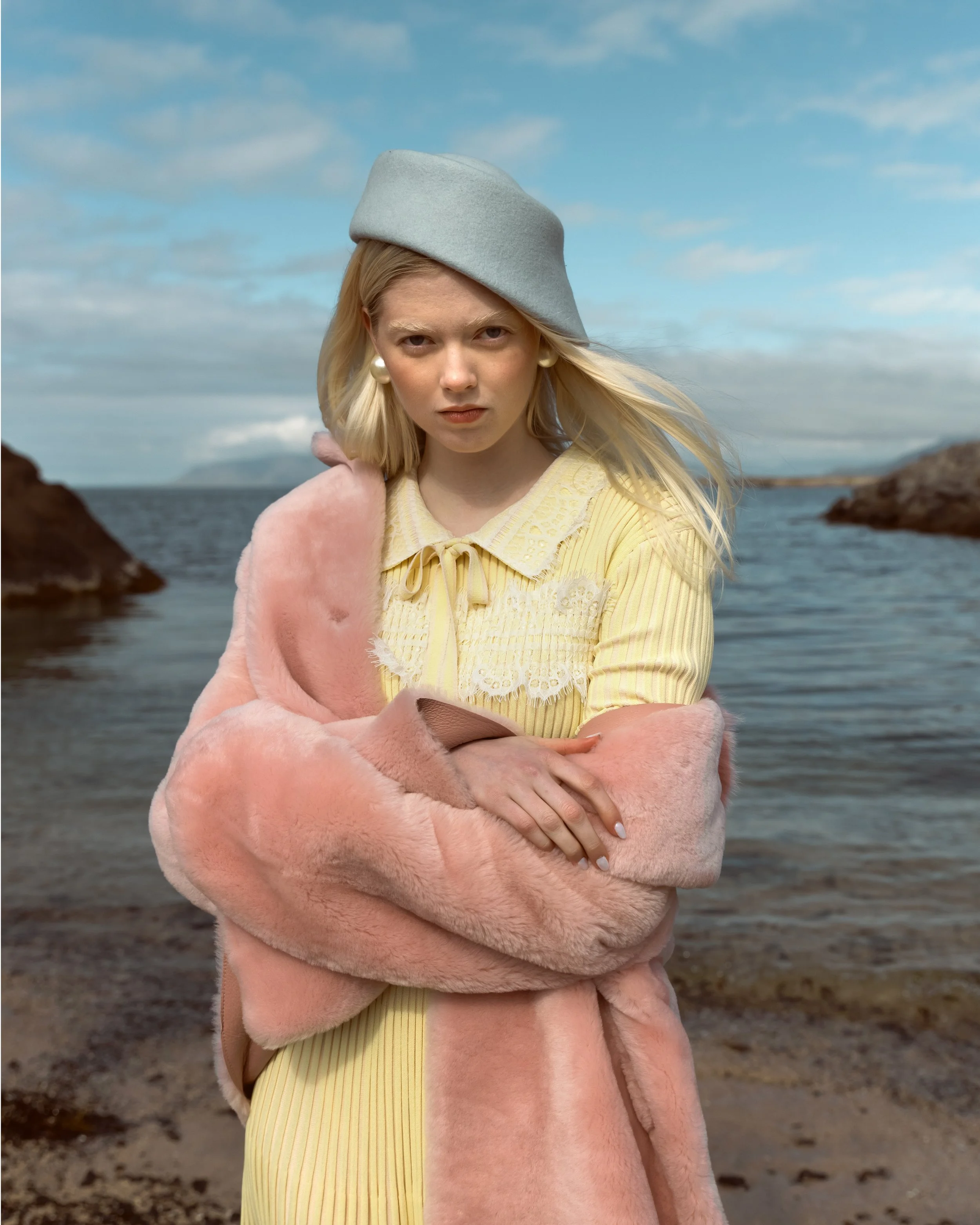 A young woman standing on a rocky beach with water and a cloudy sky in the background. She has blonde hair, wears a light gray beret, a yellow dress with lace details, and a pink fur coat. She has a serious expression and her arms are crossed.