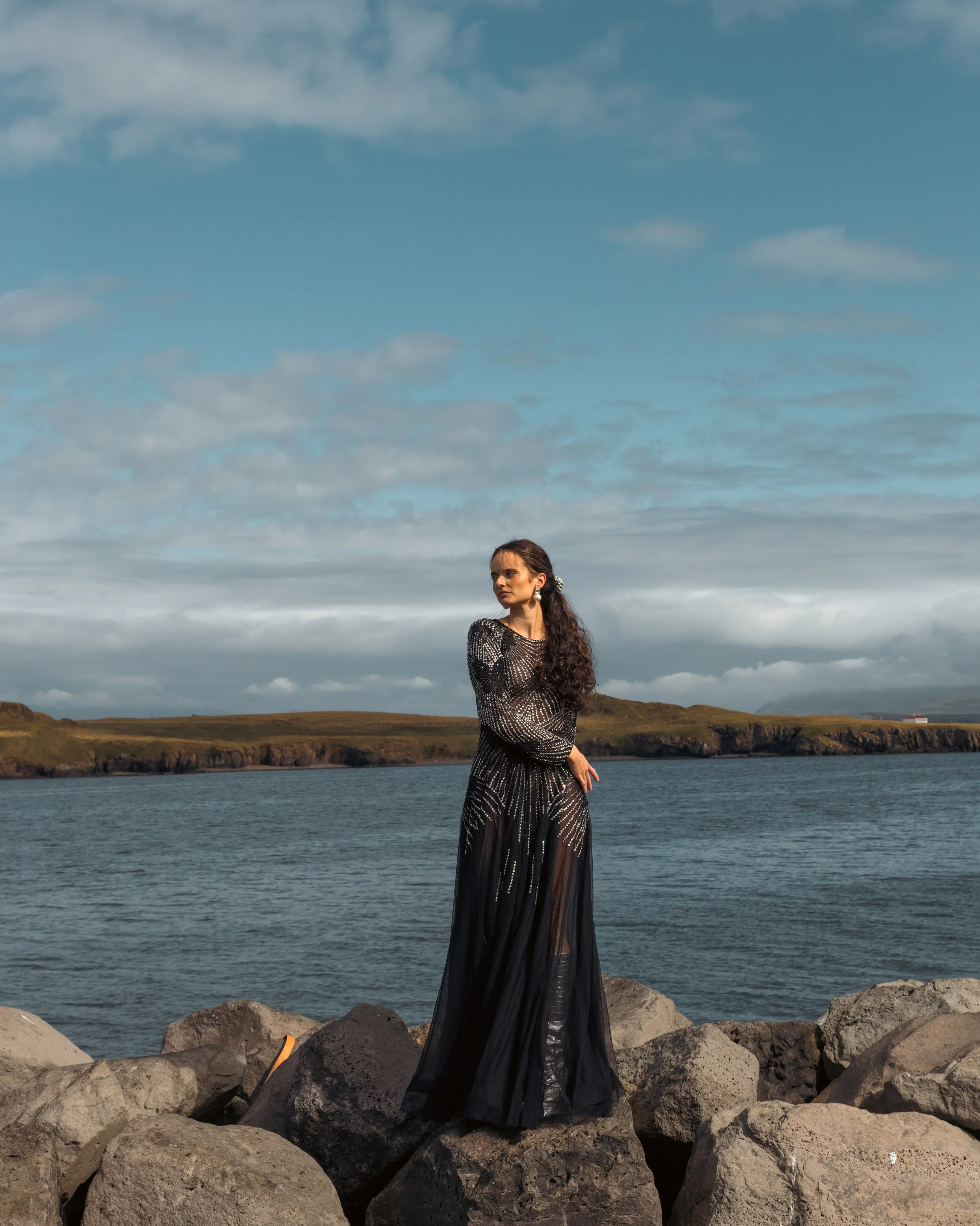A woman in a long black dress standing on rocks near a body of water with a landscape of hills and a cloudy sky in the background.