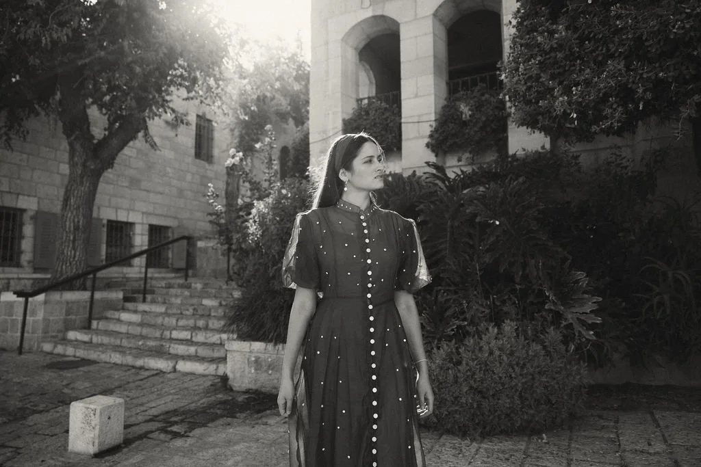 A woman in a vintage dress standing outdoors on a stone path with steps leading to a building, surrounded by trees and bushes, in black and white.