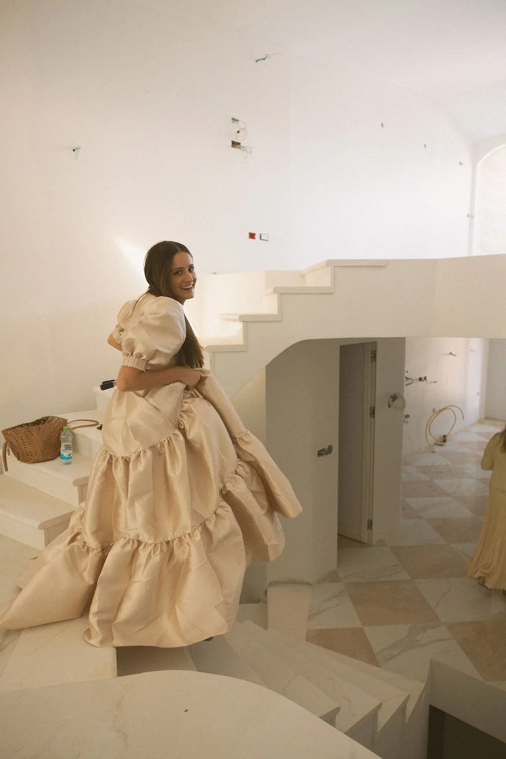 A woman wearing a beige ball gown with ruffles sitting on a white table in a mostly empty room with white walls, staircase, and marble flooring, smiling at the camera.