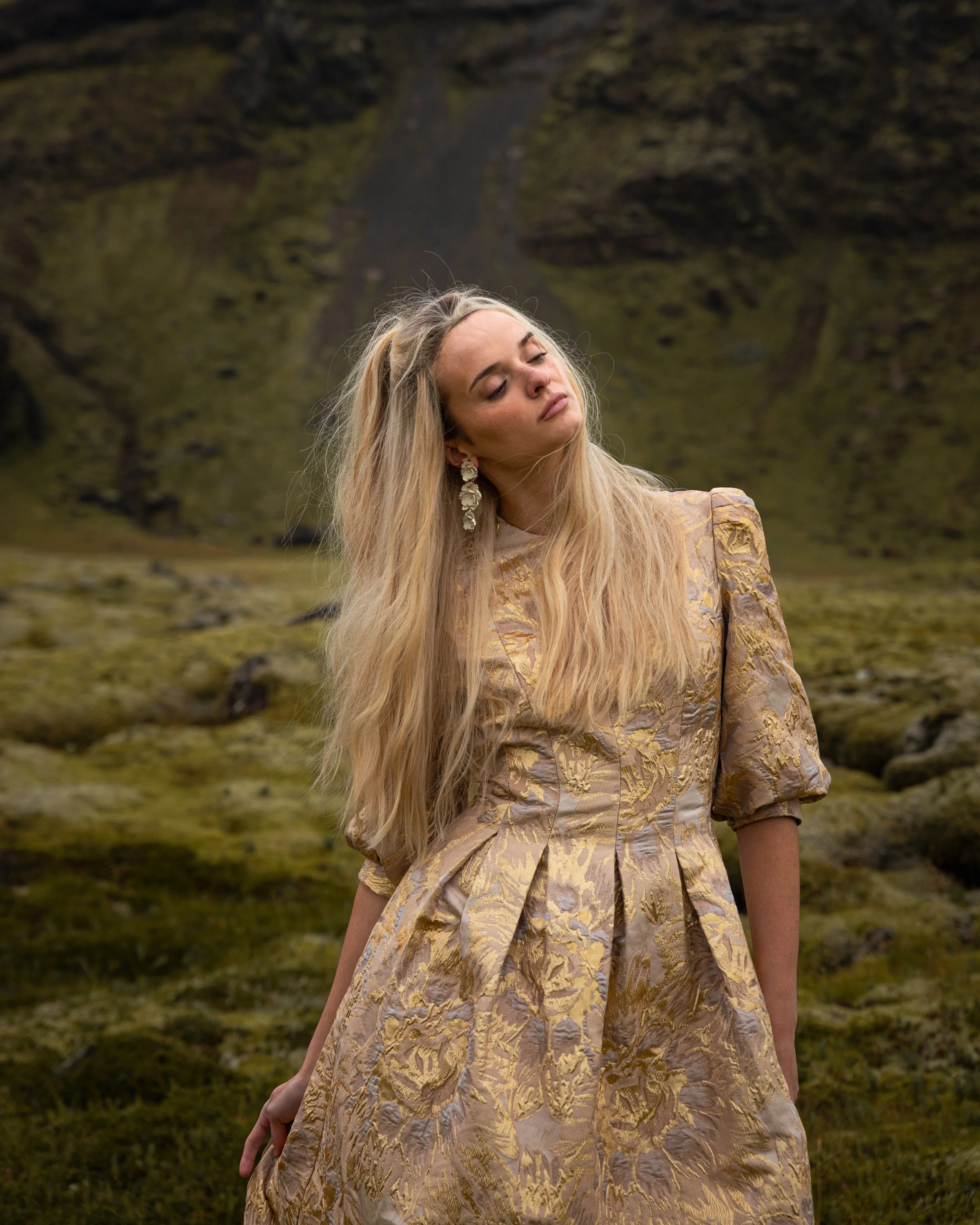 A woman with long blonde hair and earrings wearing a gold-patterned dress standing outdoors in a mossy landscape.