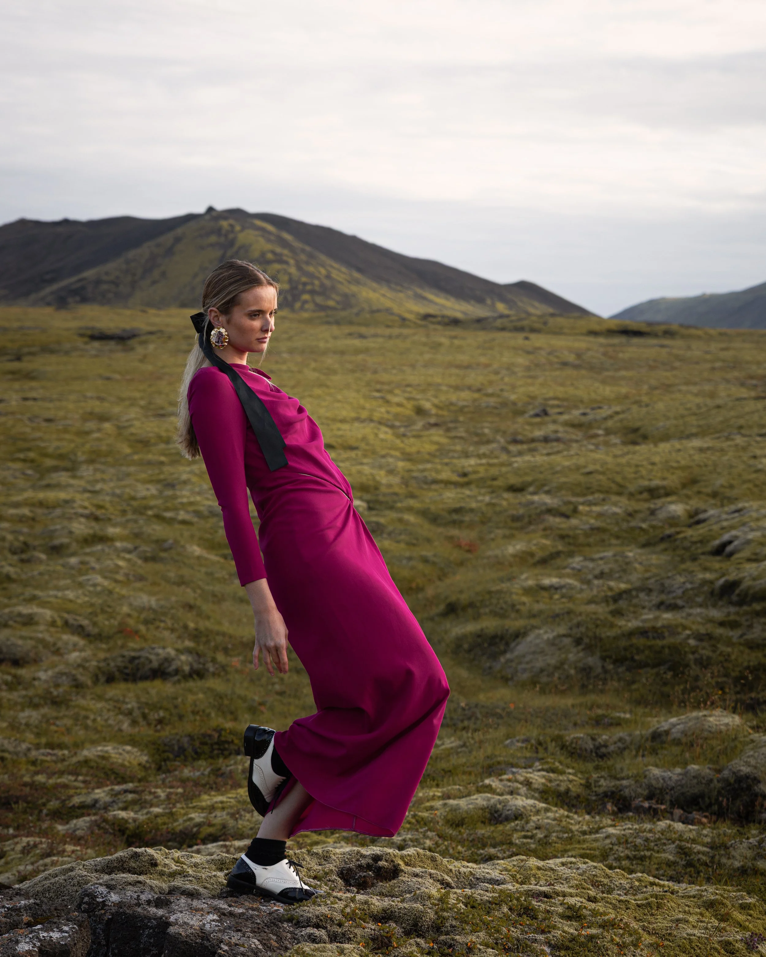 A woman in a magenta dress and black-and-white shoes standing on rocky terrain in a mountainous landscape with overcast skies.