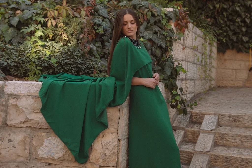 woman in a long green dress leaning against a stone wall with greenery in the background.