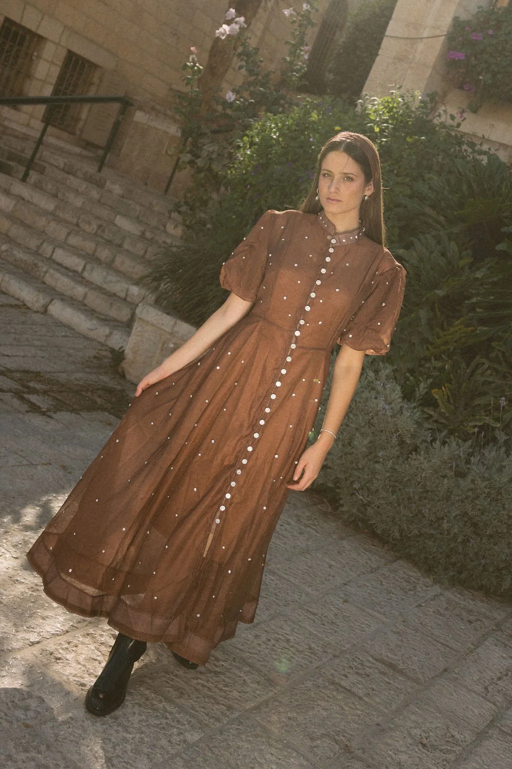 A woman in a long, brown, button-down dress with puffy sleeves, standing outdoors on a stone path, with greenery and stone buildings in the background, illuminated by sunlight.
