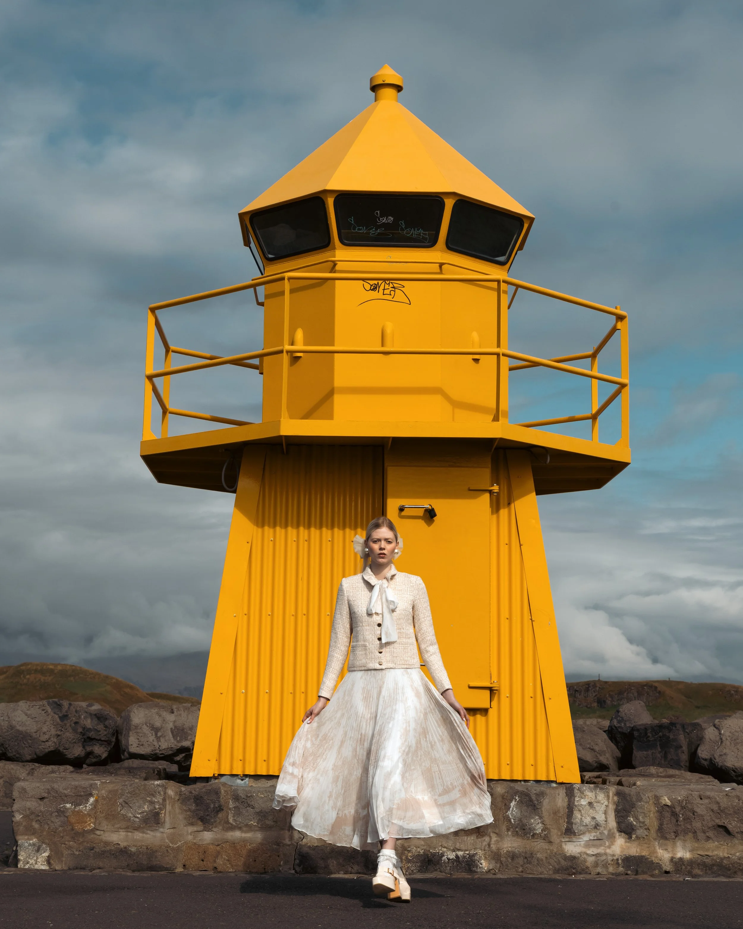 A woman in a vintage style white dress and beige blazer standing in front of a bright yellow lighthouse on a cloudy day.