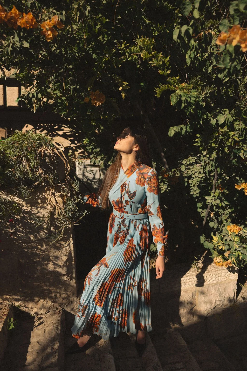 A woman in a floral dress standing in a garden with sunlight casting shadows on her face and surroundings.