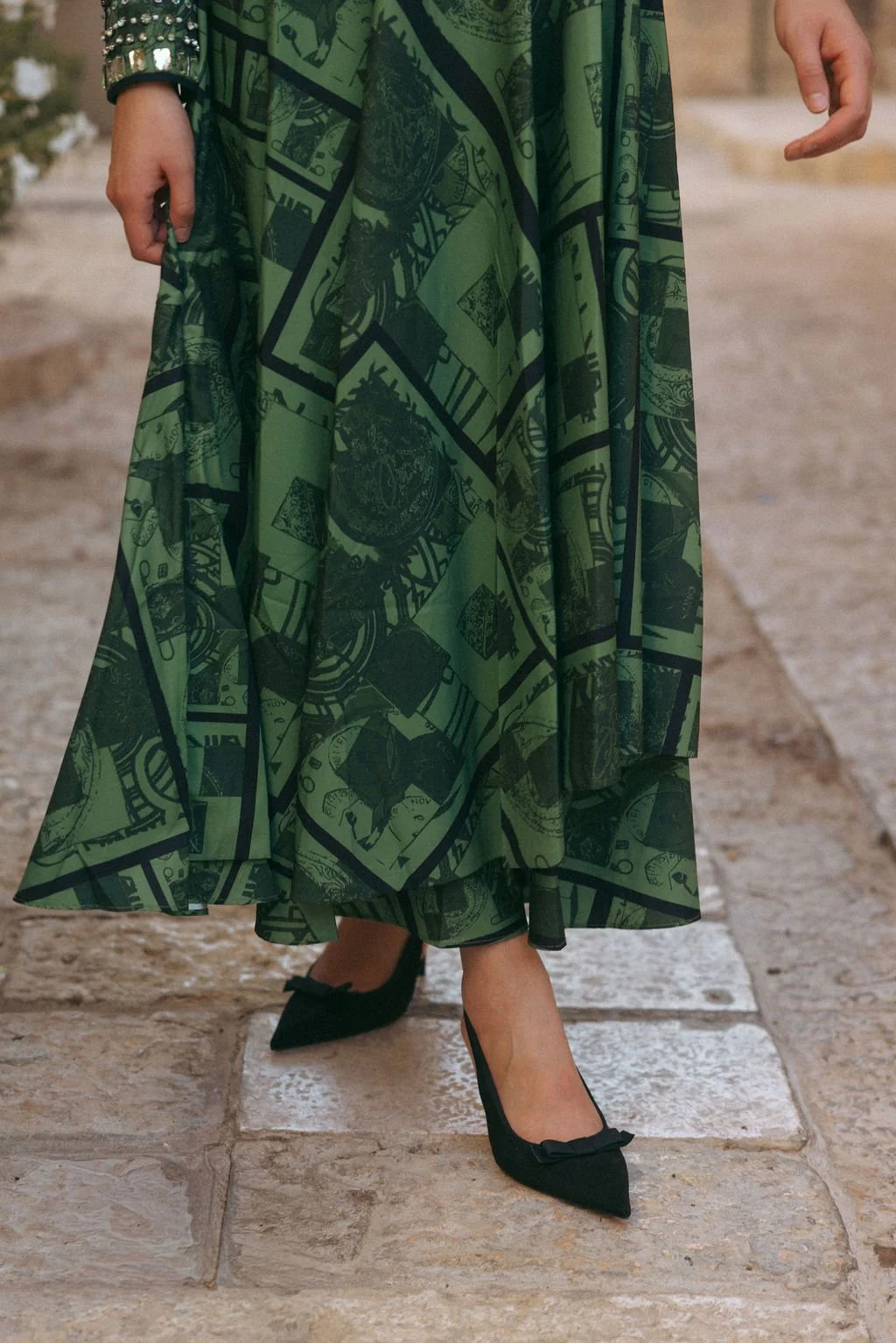 Close-up of a person wearing a long green dress with black printed patterns, standing on stone tiles, with black pointed heels featuring bows.