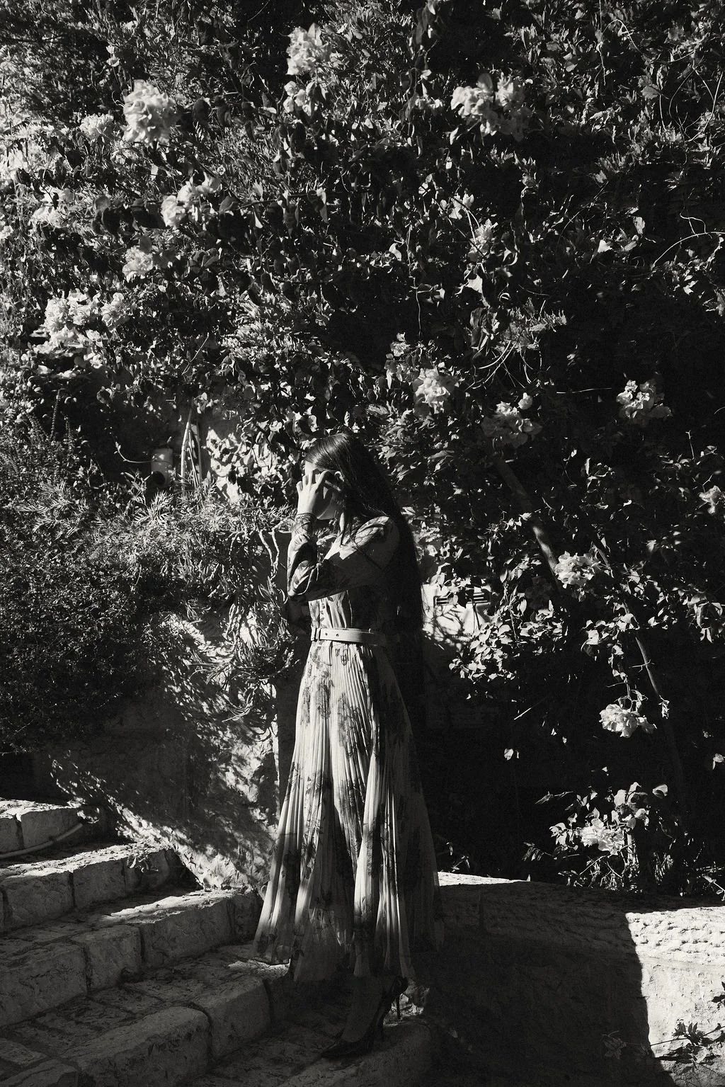 A woman standing on stone steps outdoors, surrounded by dense foliage and flowering bushes, with sunlight casting shadows.