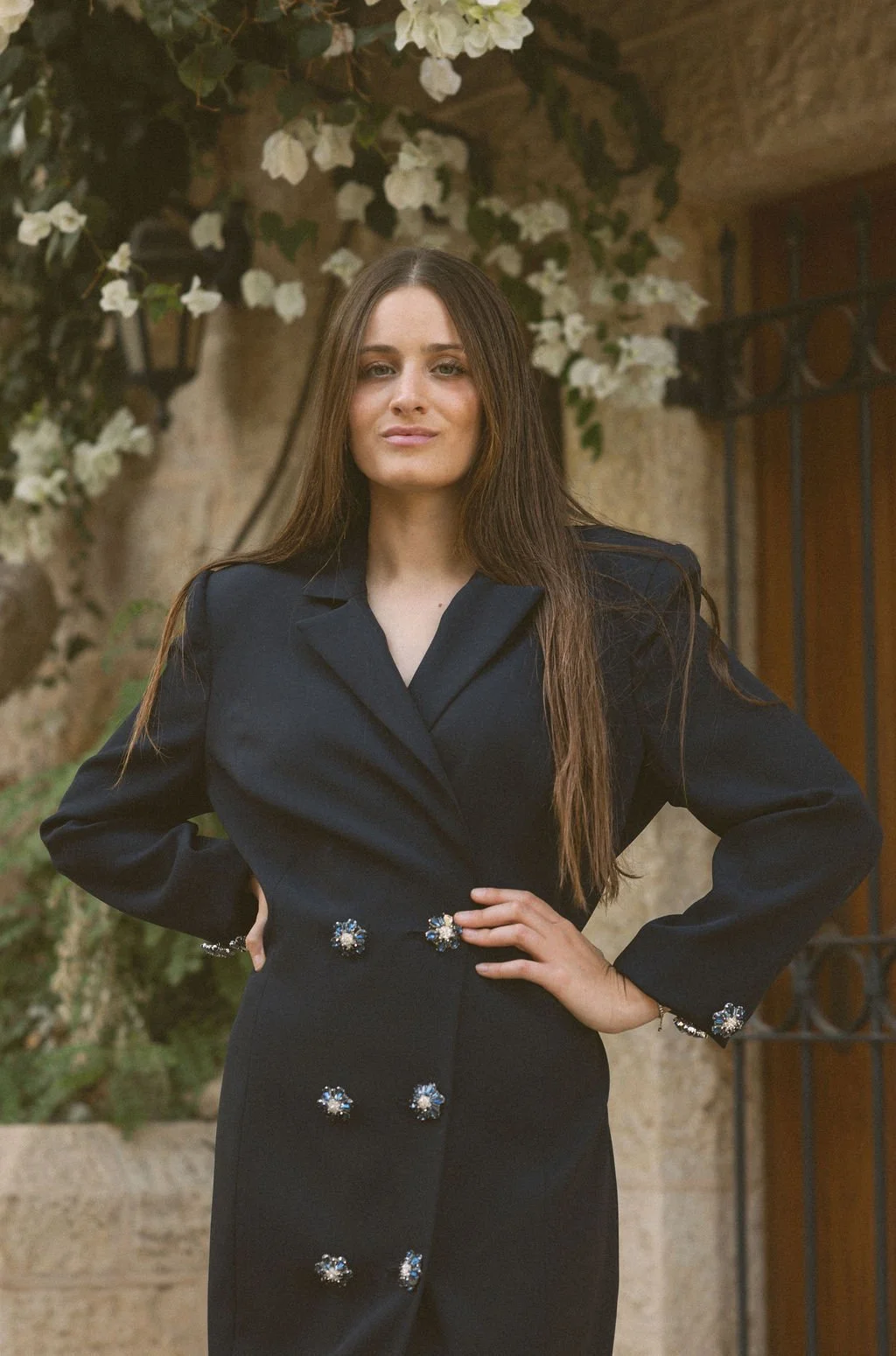 A woman with long brown hair wearing a black suit with decorative buttons stands with her hand on her hip outside in front of a stone wall and white flowering bushes.