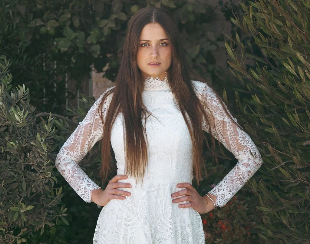 A woman with long brown hair wearing a white lace dress standing outdoors with greenery and bushes in the background.
