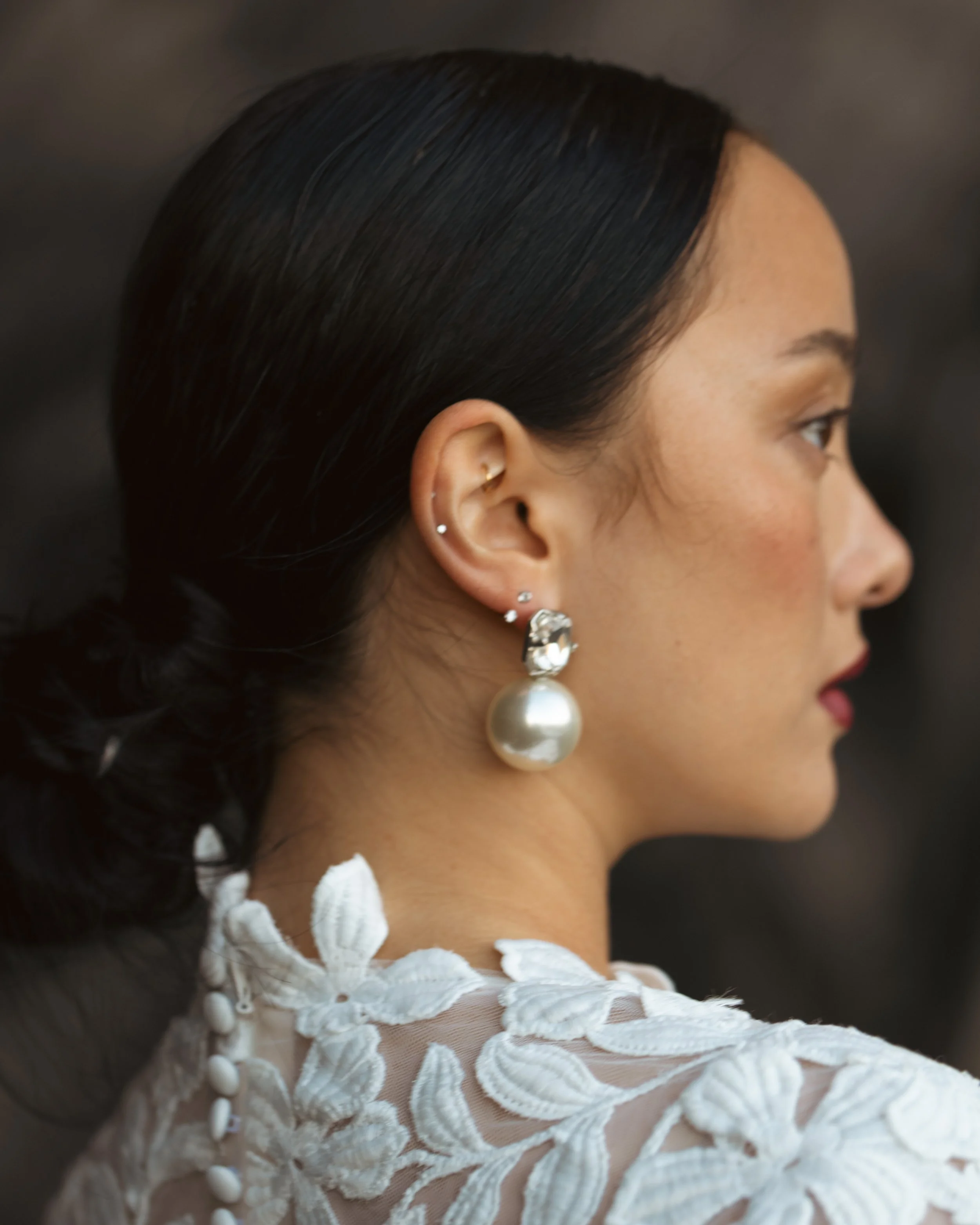 A profile view of a woman with dark hair styled in a low bun, wearing large pearl and diamond earrings, and a white embroidered lace dress.