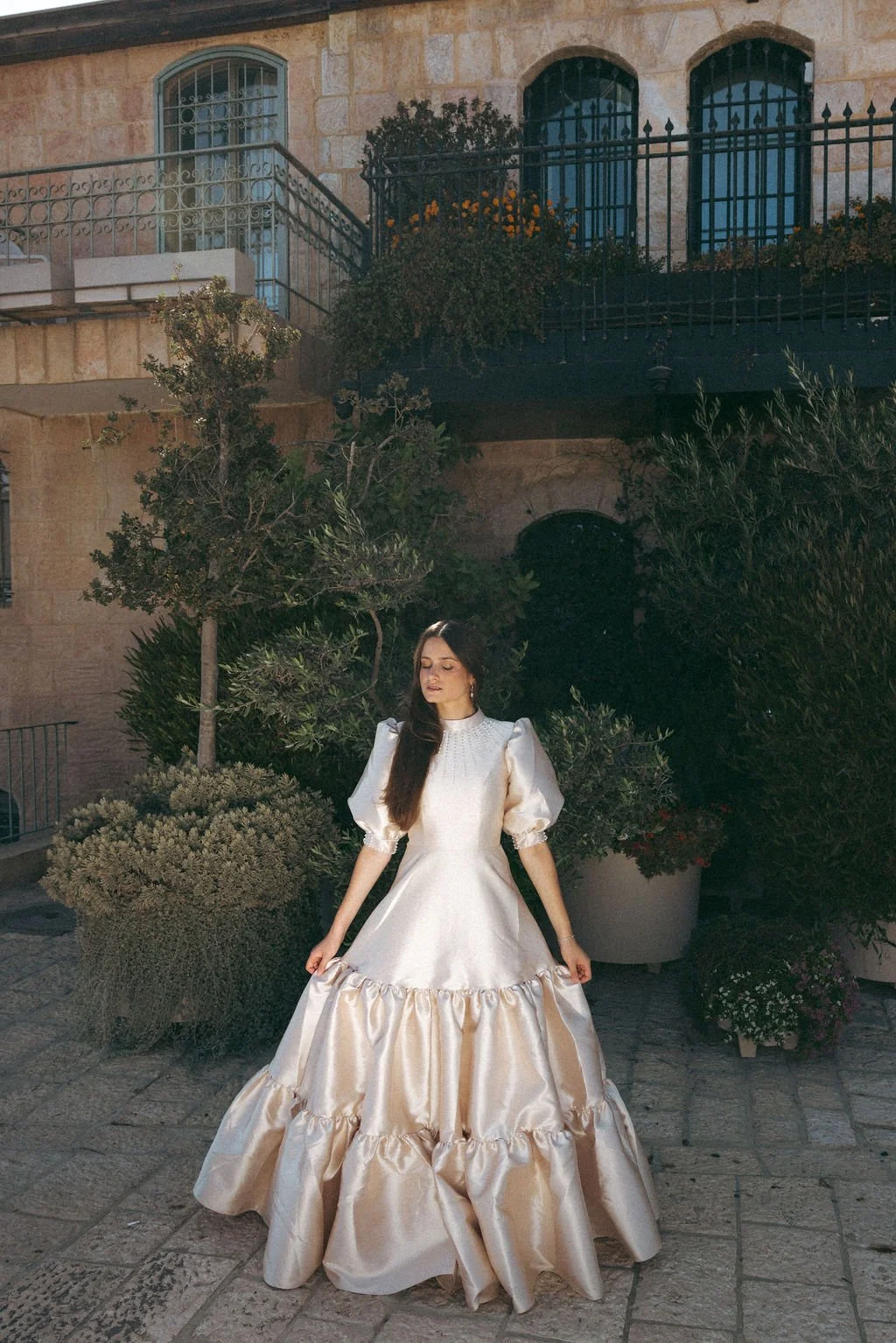 A woman in an elegant, vintage-style white satin dress with puffed sleeves and ruffled skirt, standing outdoors at dusk with large potted plants and an old stone building in the background.