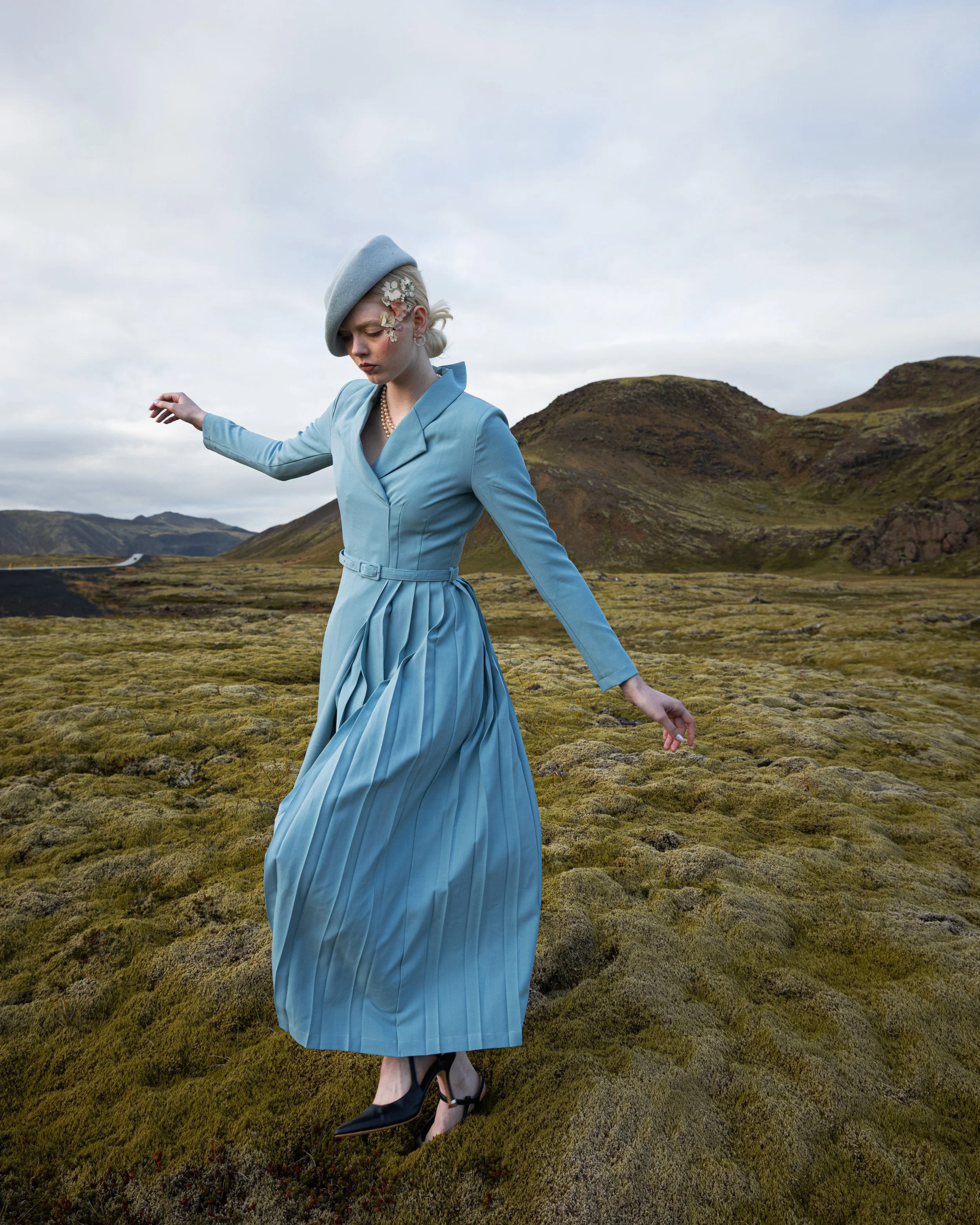 A woman in a blue dress and gray beret walking on mossy ground in a mountainous landscape.