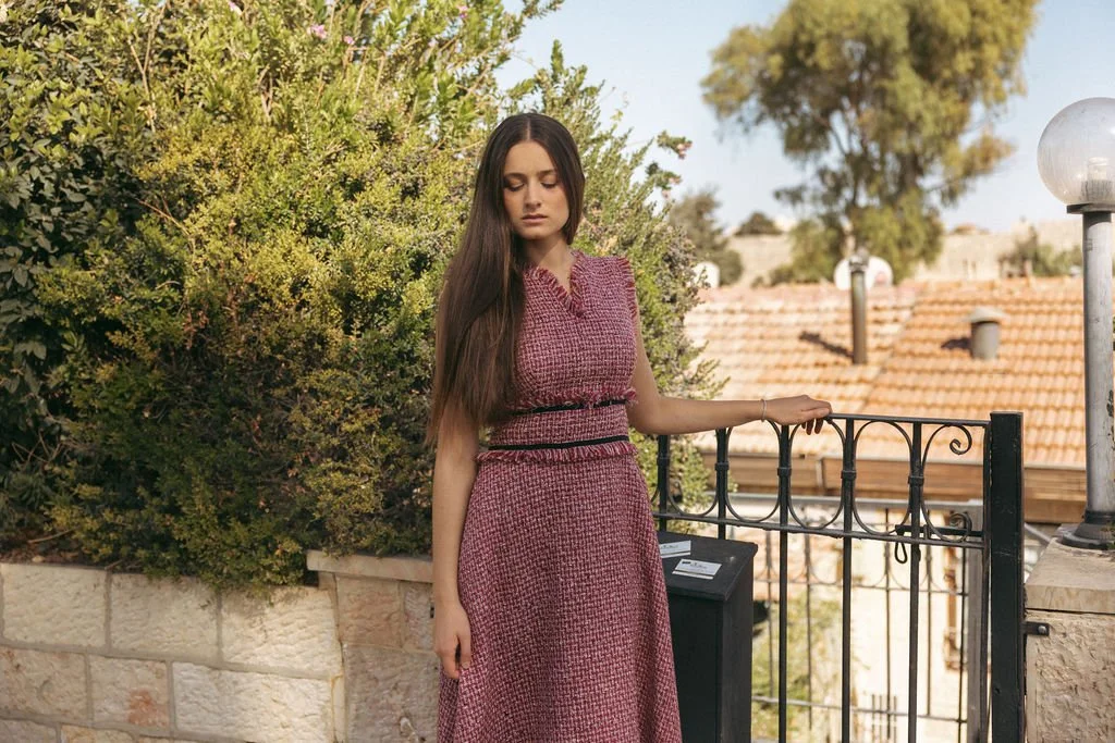 A young woman with long brown hair wearing a matching sleeveless pink textured dress standing outdoors near a black fence, with trees and rooftops visible in the background.