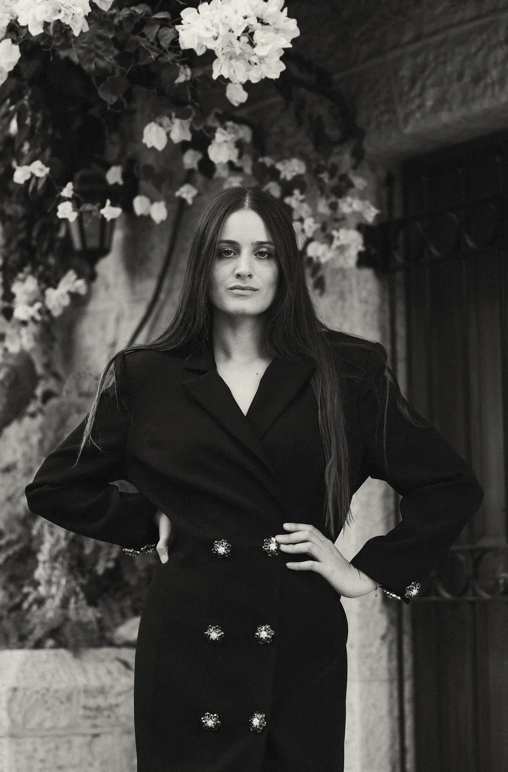 A woman with long dark hair wearing a dark suit with decorative buttons, standing with hands on hips in front of a background with flowers and a stone wall.