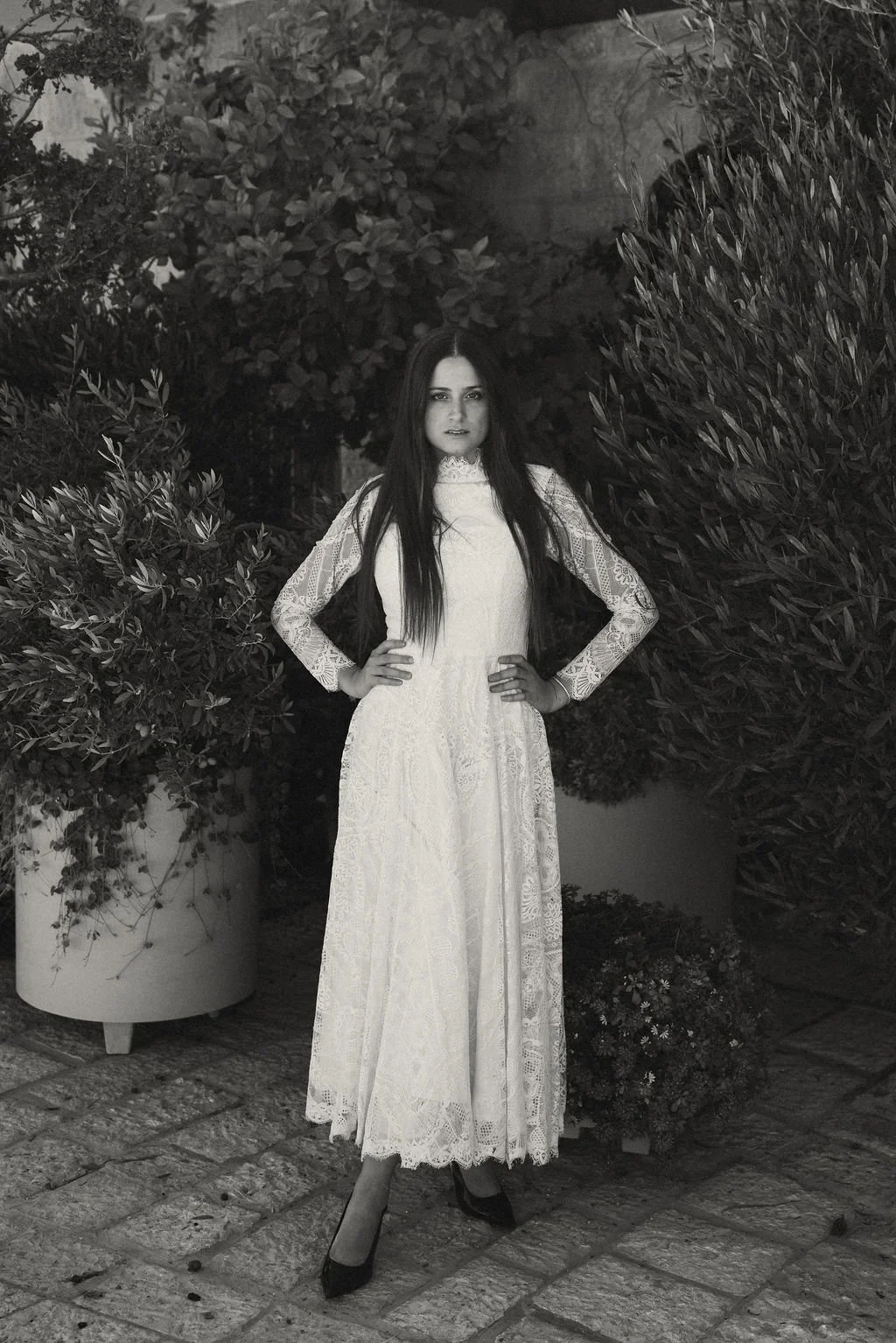 A woman in a long lace dress standing outdoors on a stone patio, surrounded by lush bushes and trees.