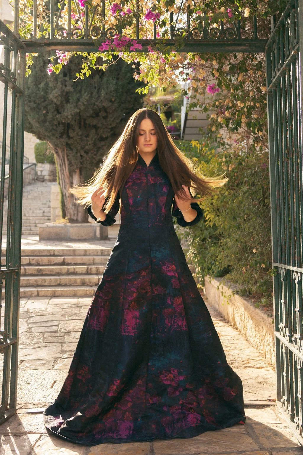 A woman in a long, dark floral dress walking through an open wrought iron gate in a garden, with stairs and trees in the background, illuminated by warm sunlight.