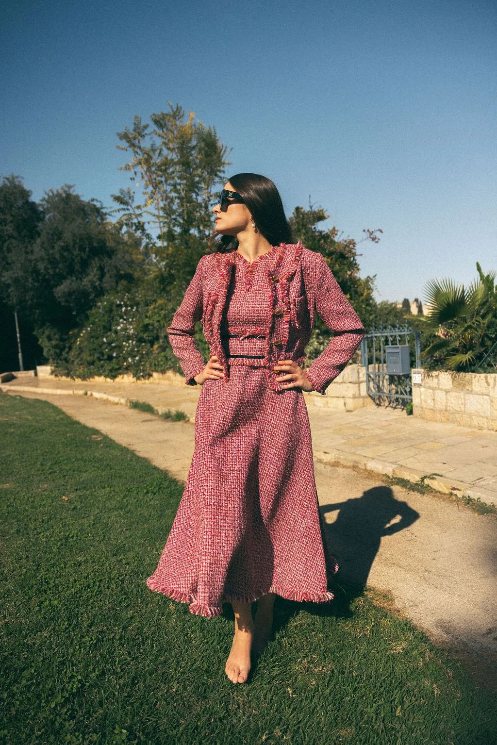 A woman in a pink patterned suit, wearing sunglasses, standing outdoors on a sunny day with green grass, trees, and a stone wall in the background.