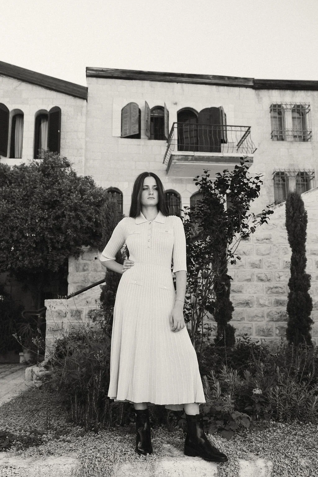 Black and white photo of a woman standing outdoors in front of a stone house with shuttered windows, a small balcony, and lush plants and trees.