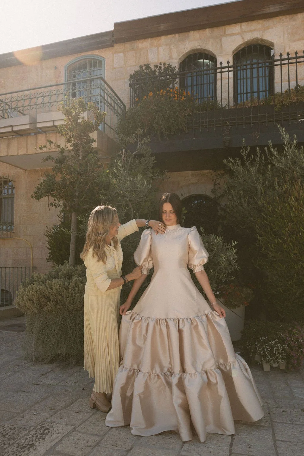 Two women, one helping the other with a white gown, standing outdoors in front of a stone building with balconies and greenery.